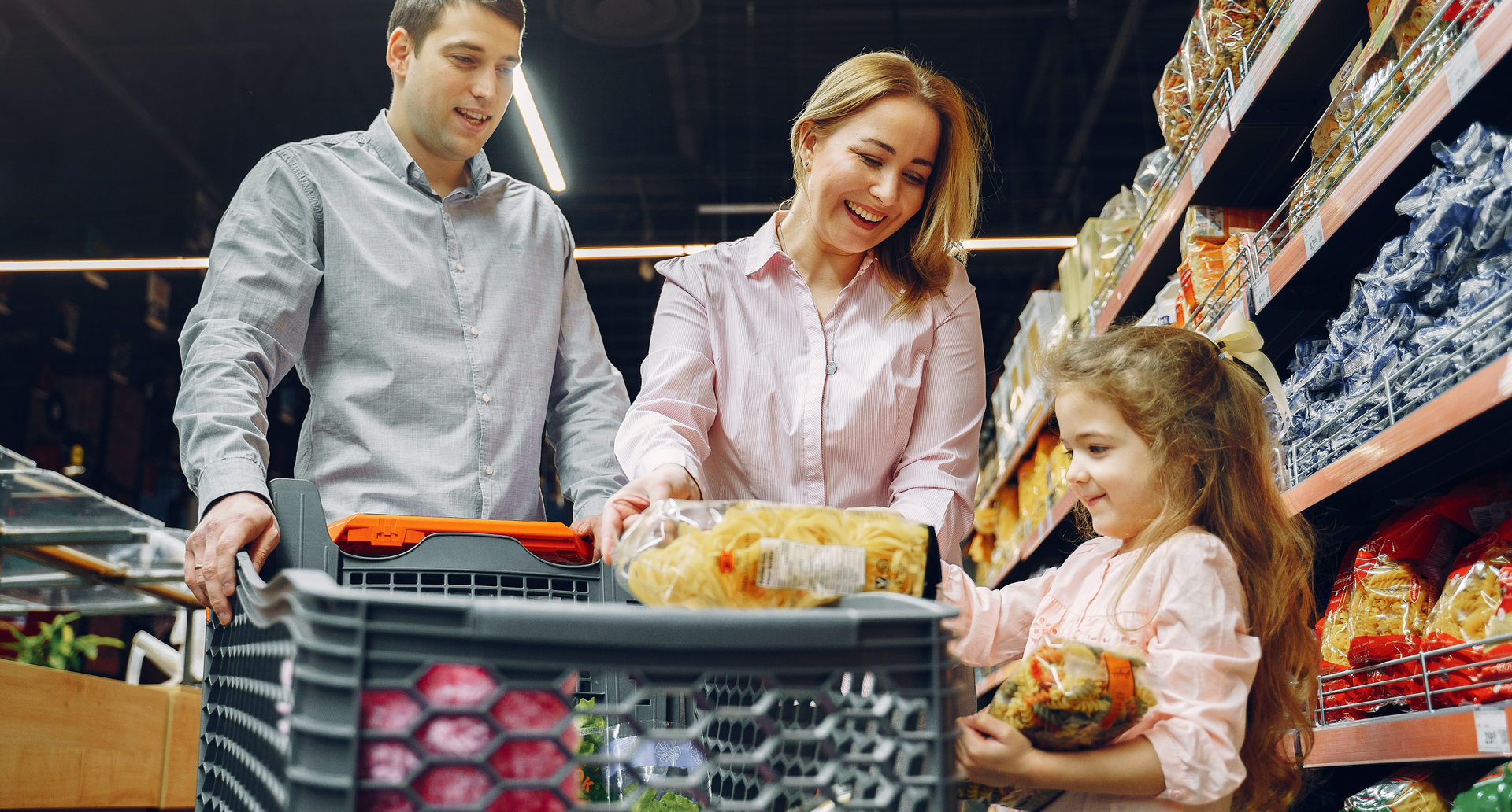 Zdjęcie przedstawia mężczyznę, kobietę i dziewczynkę robiących zakupy w sklepie. Są uśmiechnięci, mają ze sobą wózek na zakupy. Dziewczynka włada produkty do wózka.