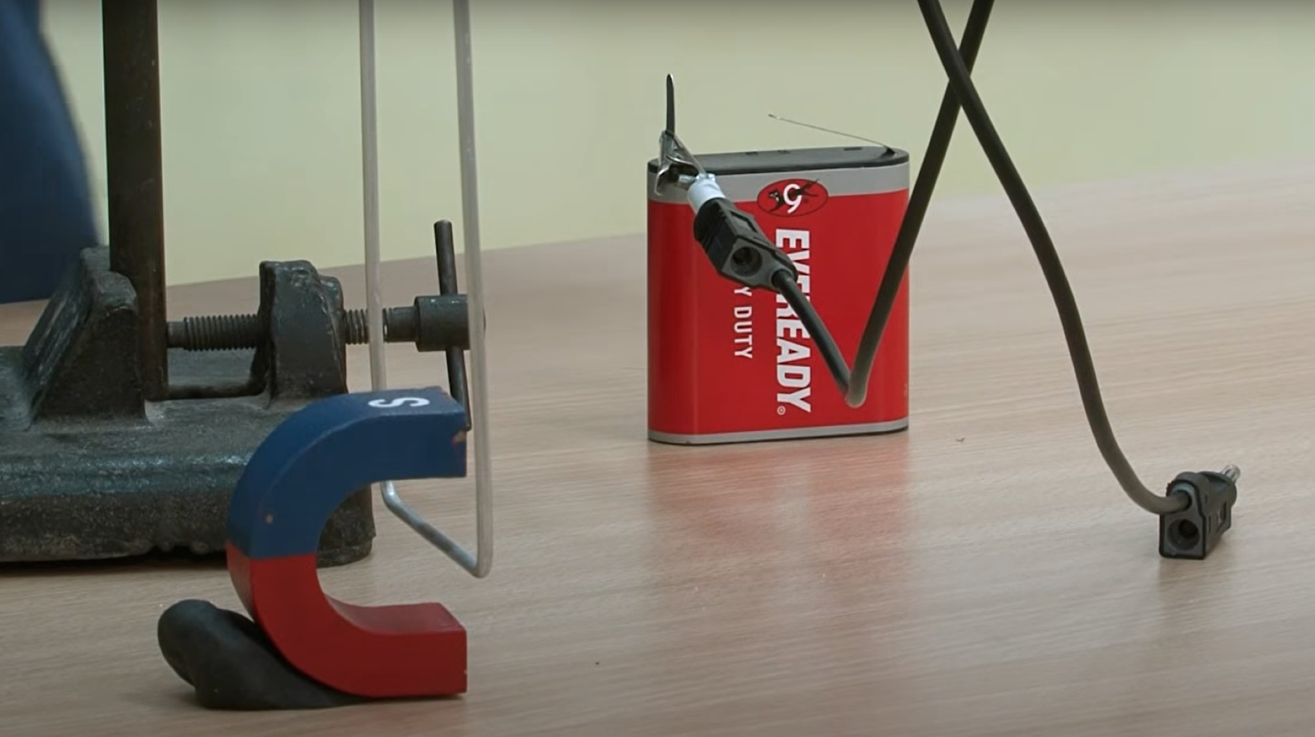 Rys. 3. Zdjęcie przedstawia pionowy statyw, do którego górnej części przymocowano poziomy, biały uchwyt w kształcie walca. Na uchwycie zawieszono huśtawkę, składającą się z dwóch pionowych aluminiowych drutów połączonych w dolnej części aluminiową poprzeczką. Poprzeczkę umieszczono między biegunami magnesu w kształcie podkowy. Magnes ustawiony jest tak, że jego biegun wielka litera N, pomalowany na czerwono, jest nad poprzeczką, a biegun S pomalowany na niebiesko jest pod poprzeczką.