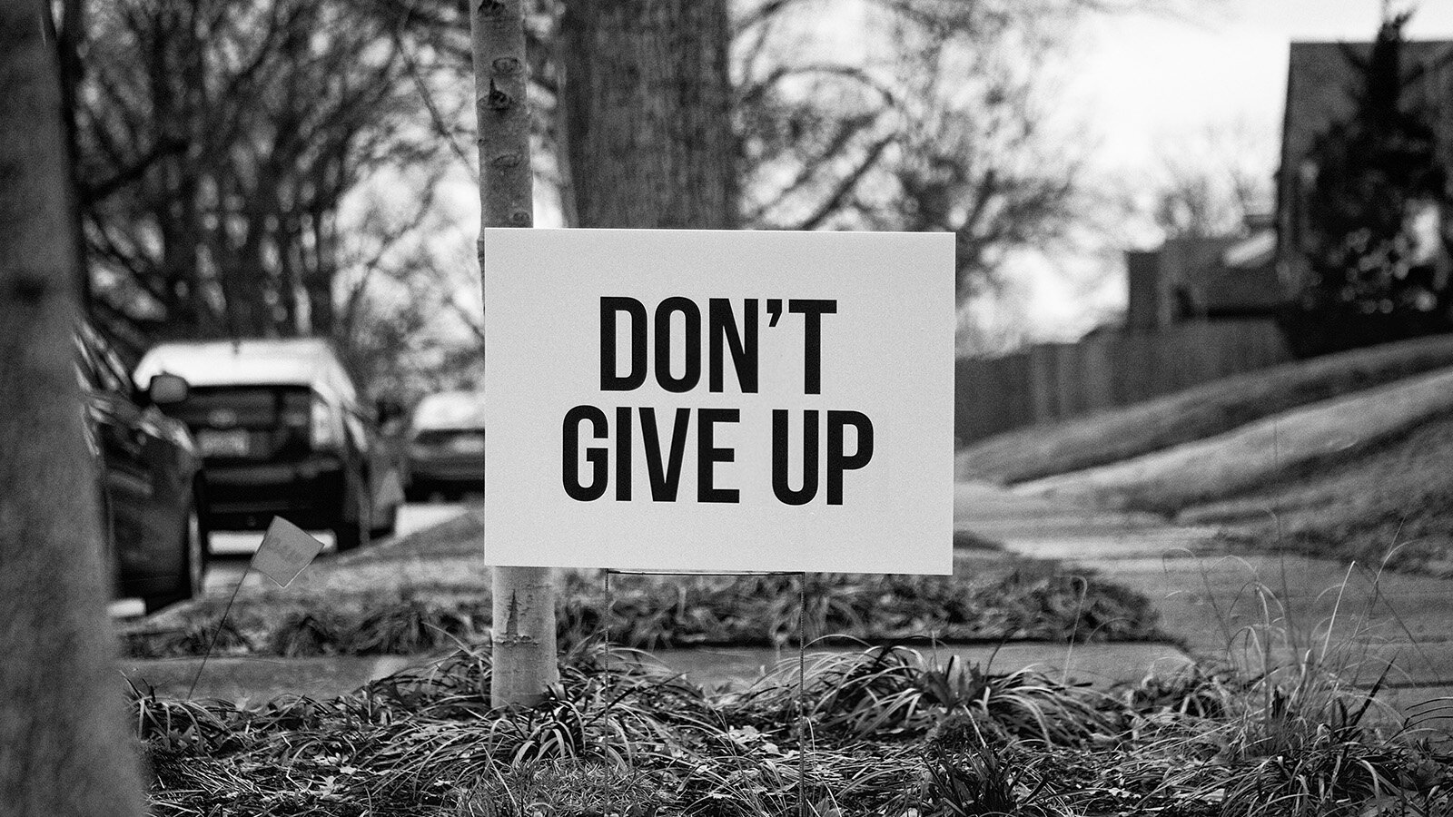 Czarno‑białe zdjęcie przedstawia fragment ulicy z widocznymi po lewej stronie zaparkowanymi samochodami, pasem zieleni pośrodku i chodnikiem po prawej stronie. Pośrodku zdjęcia widoczna biała tabliczka z napisem w czarnym kolorze w języku angielskim: DON'T GIVE UP.
