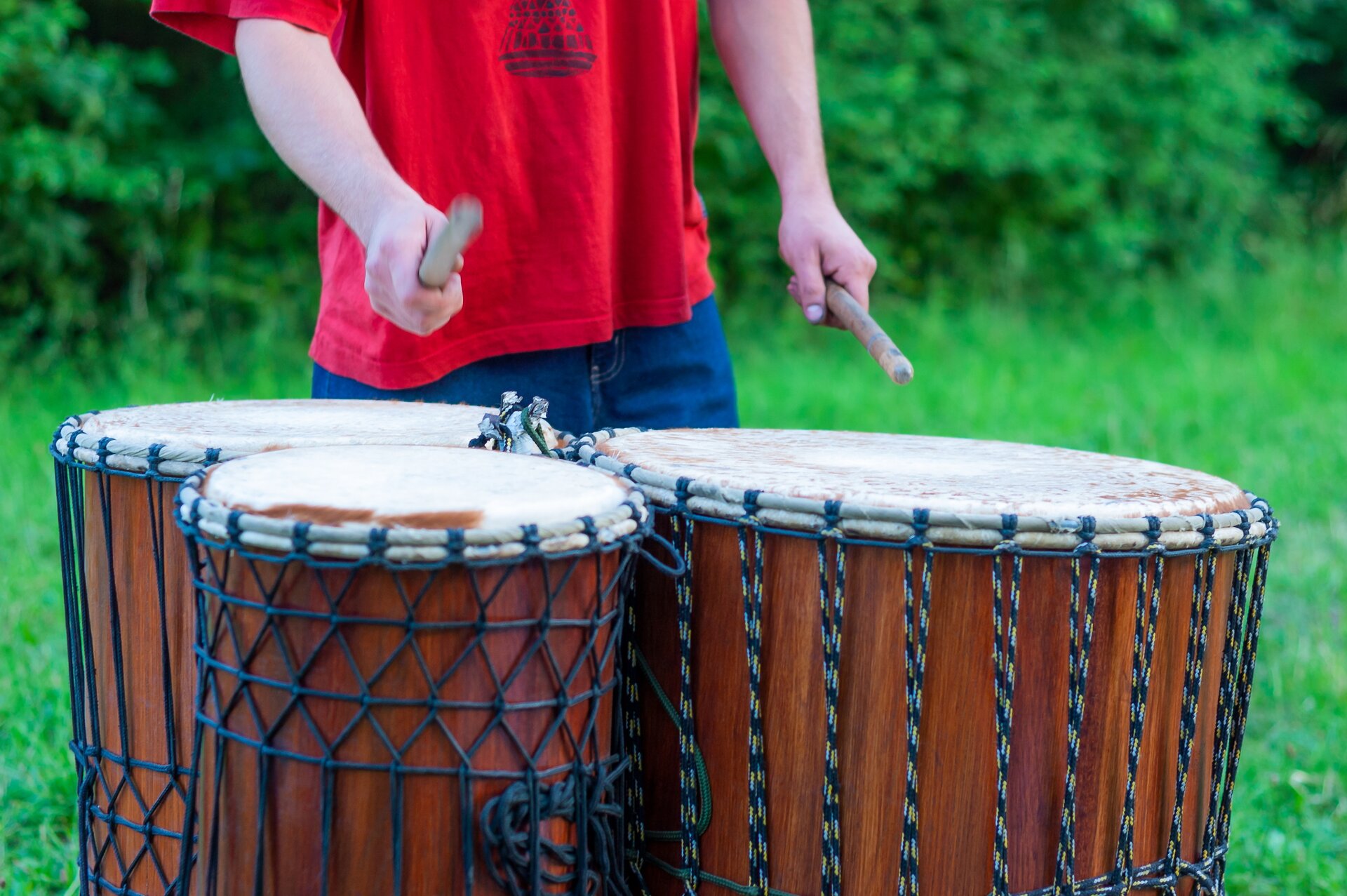 Na pierwszym planie obecne są trzy duże przedmioty przypominające brązowe walce, których boki oplecione czarnym sznurkiem, a wierzch – kremowym materiałem. Są to dunduny - instrumenty perkusyjne pochodzące z Afryki. Mają one równą wysokość, ale różnią się średnicą. Za bębnami znajduje się sylwetka postaci ubranej w niebieskie spodnie oraz czerwoną koszulkę z czarną grafiką, trzymającej w obu dłoniach krótkie, grube pałeczki do gry. Tło stanowi zielona trawa oraz liście krzewów.