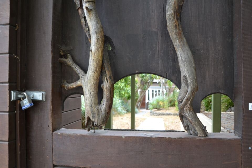 Ilustracja przedstawia fotografię fragmentu bramy wejściowej na posesję miejską. Brama posiada trzy półkoliście zakończone otwory, które znajdują się w środkowej części bramy. Widać przez nie fragment drogi oraz budynku. Na bramie znajdują się konary drzew, pozbawione liści - są one elementem dekoracji bramy.