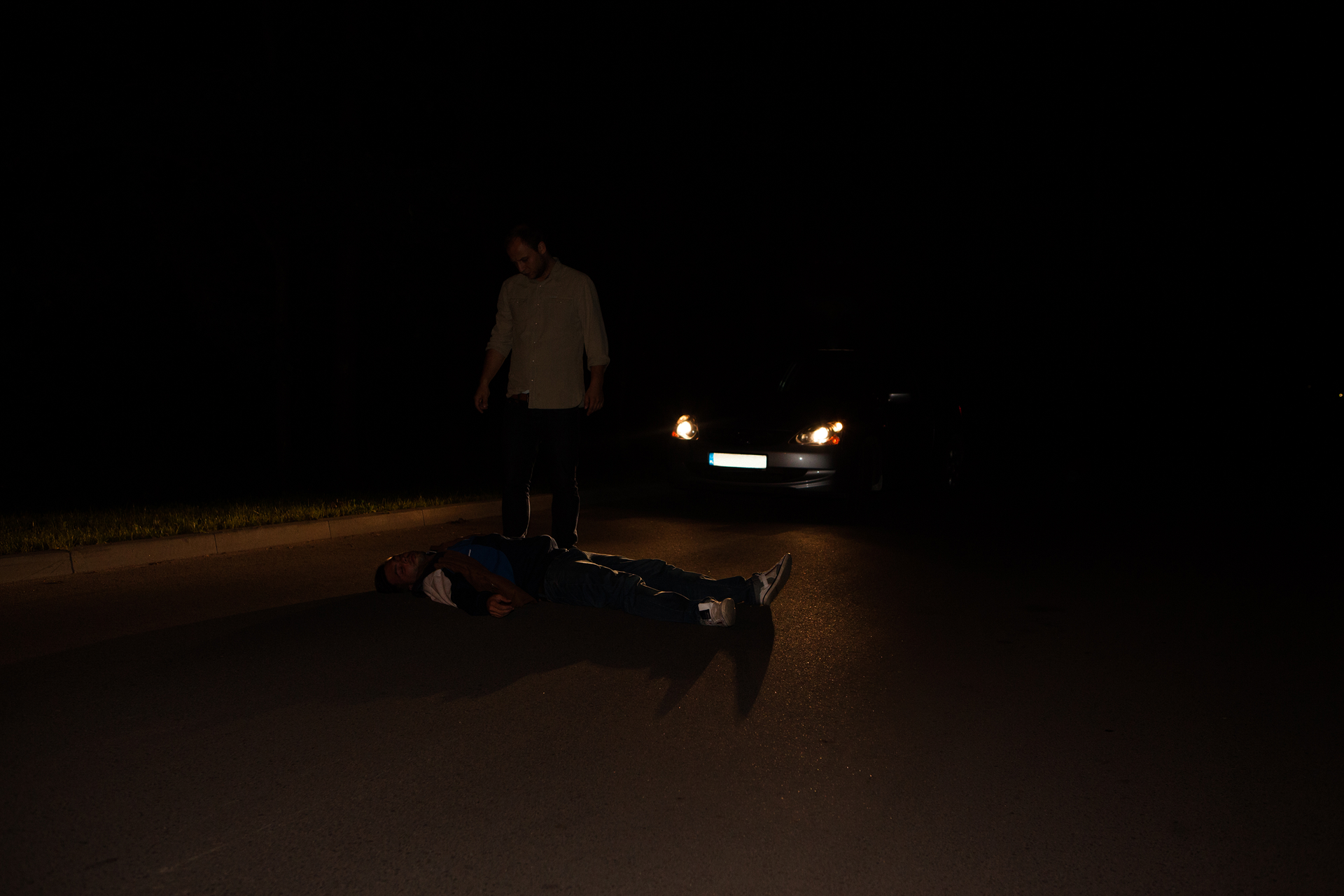 Noc. Na jezdni leży nieprzytomna osoba. Leżąca osoba bez odblaskowych dodatków. Za leżącą osobą zaparkowany samochód z włączonymi reflektorami. Reflektory skierowane na leżącą osobę. Kierowca bez kamizelki odblaskowej jest prawie niewidoczny na jezdni.