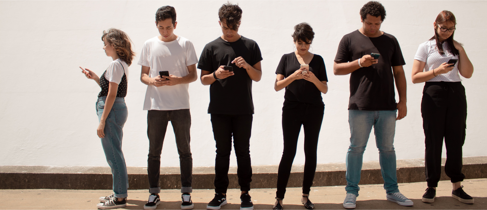 Zdjęcie przedstawia sześcioro młodych ludzi, trzy dziewczyny i trzech chłopaków. Po lewej stronie znajduje się dziewczyna, która stoi bokiem do kadru i tyłem do reszty osób. Dalej przodem do kadru stoją w kolejności: dwóch chłopaków, dziewczyna, chłopak i dziewczyna.  Widoczne są całe sylwetki młodzieży. Wszyscy wpatrzeni są w swoje smartfony.