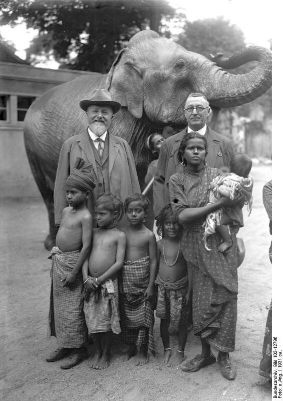 W centralnej części zdjęcia widać czwórkę indyjskich dzieci i hinduską kobietę, noszącą na rękach niemowlę. Dzieci i kobieta są przepasane tradycyjnymi strojami, kobieta i najstarsze dziecko noszą skórzane łapcie na nogach. Za nimi dwóch mężczyzn ubranych w płaszcze, garnitury i krawaty, patrzą na wprost widza. Mężczyzna po lewej nosi jasną brodę i kapelusz, mężczyzna po prawej okrągłe okulary, bez nakrycia głowy. W tle słoń indyjski prowadzony przez osobę częściowo zasłoniętą postaciami pierwszoplanowymi. Na dalszym tle zabudowania. Zdjęcie ma charakter pozowany. 