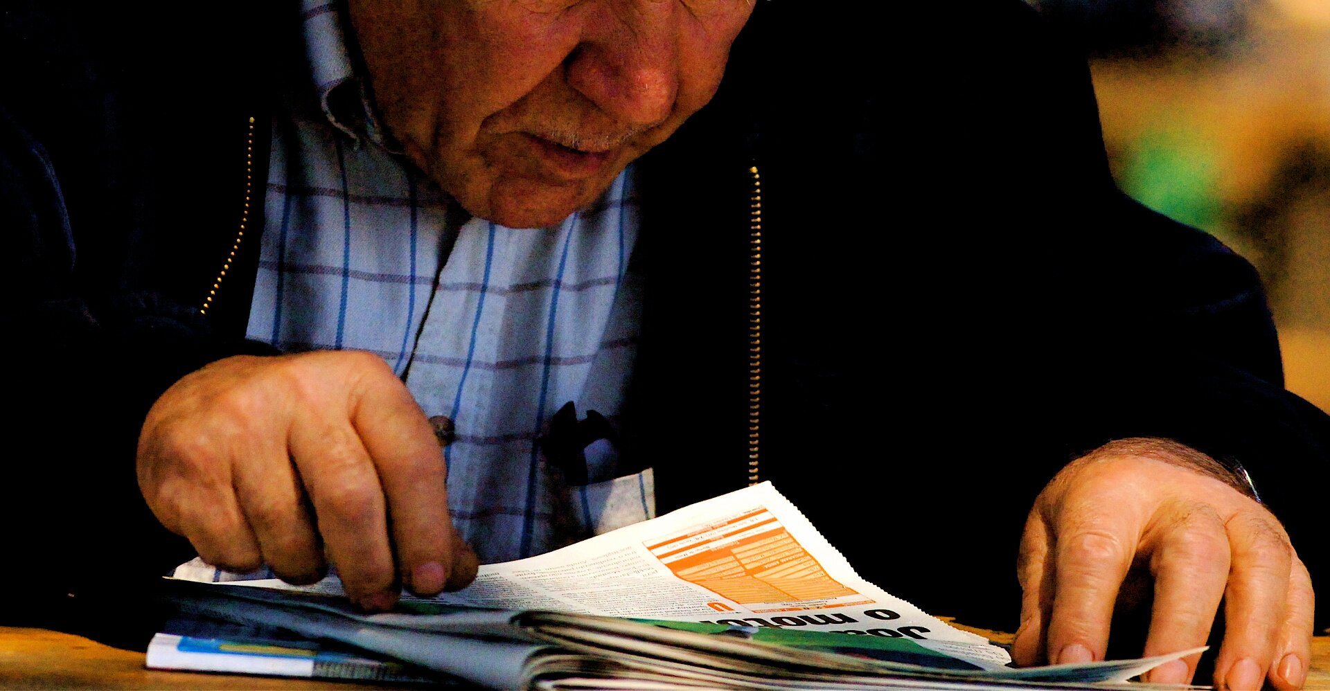 Zdjęcie przedstawia starszego mężczyznę, ubranego w koszulę w kratkę, czytającego gazetę. Jest on nad nią pochylony.