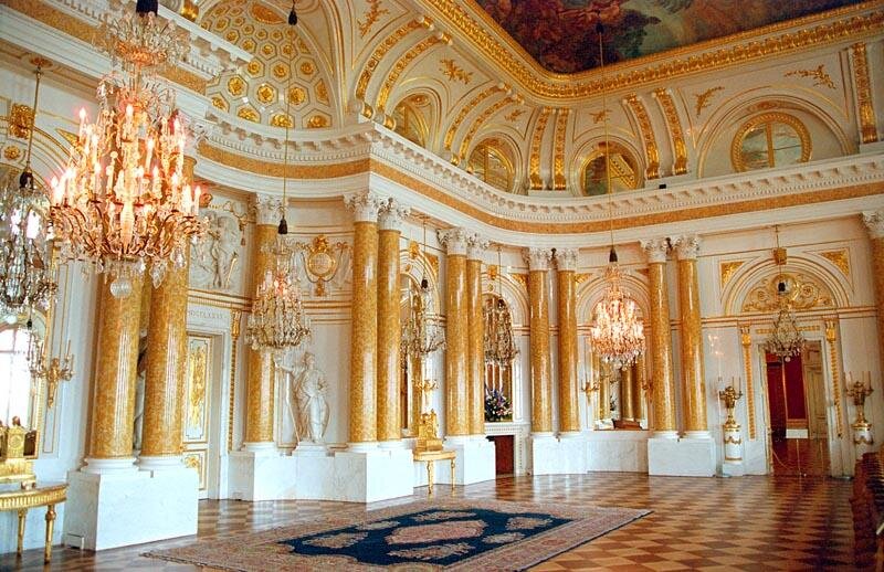 Ilustracja przedstawia salę pałacową. Wnętrze zdobią rozmieszczone koliście złotawe kolumny ze stiuku, okazałe kandelabry, żyrandole i bogato złocone dekoracje ścienne. Na suficie malowidło. Posadzka wyłożona kaflami w dwóch kolorach ułożonymi naprzemiennie w karo.
