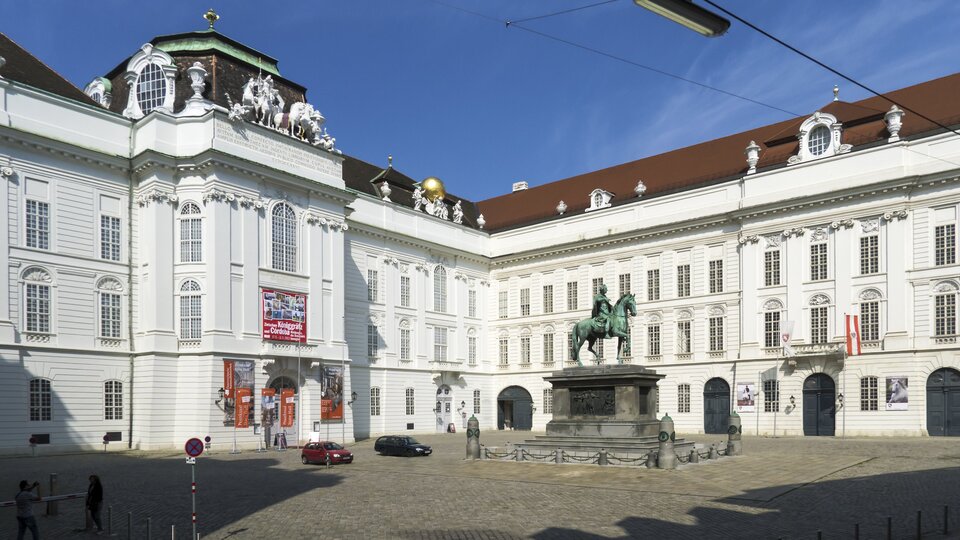 Ilustracja przedstawia budynek zaprojektowany przez Johanna Bernharda Fischer von Erlach i Joseph Emmanuel Fischer von Erlach, fasadę Biblioteki Dworskiej w Wiedniu. Trójkondygnacyjna fasada pokryta jest białą elewacją, środkowy ryzalit jest lekko wysunięty do przodu. Na gzymsie ustawione są rzeźby.