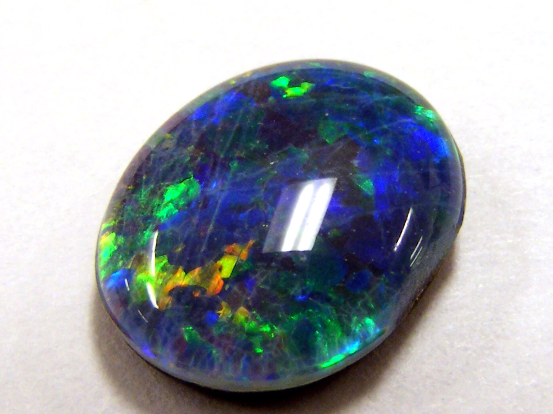 Zdjęcie przedstawia oszlifowany opal niebieski w odmianie szlachetnej, czyli opalizującej, co oznacza zmienność barw w zależności od kąta obserwacji. Szlif owalny, kamień ma formę nieco spłaszczonej kropli.