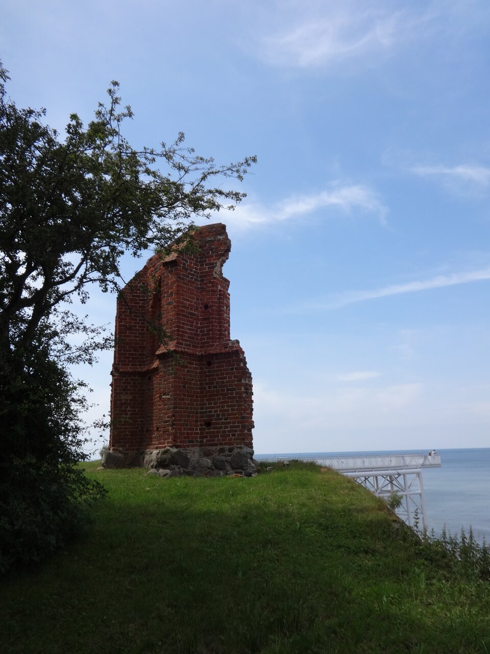 Zdjęcie ruin kościoła w Trzęsaczu z 2014 r. Obraz przedstawia gruzy w wyglądzie, jaki mają współcześnie. W tle wysunięty, wsparty na palach taras widokowy i morze. 