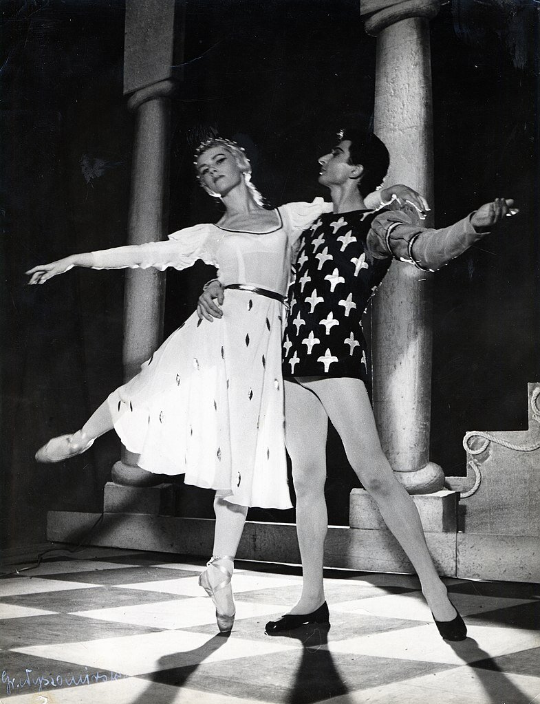 Czarno‑białe zdjęcie przedstawia scenę baletową. Na tle dwóch kolumn stoją dwie postacie: kobieta i mężczyzna. Kobieta w białej sukience stoi na palcach lewej nogi, prawą ma uniesioną do góry. Trzyma za ramię mężczyznę w rajtuzach i kaftanie zdobionym krzyżykami. Oboje mają uniesione ręce: kobieta prawą, mężczyzna lewą. Kobieta patrzy w dal, mężczyzna patrzy na nią.