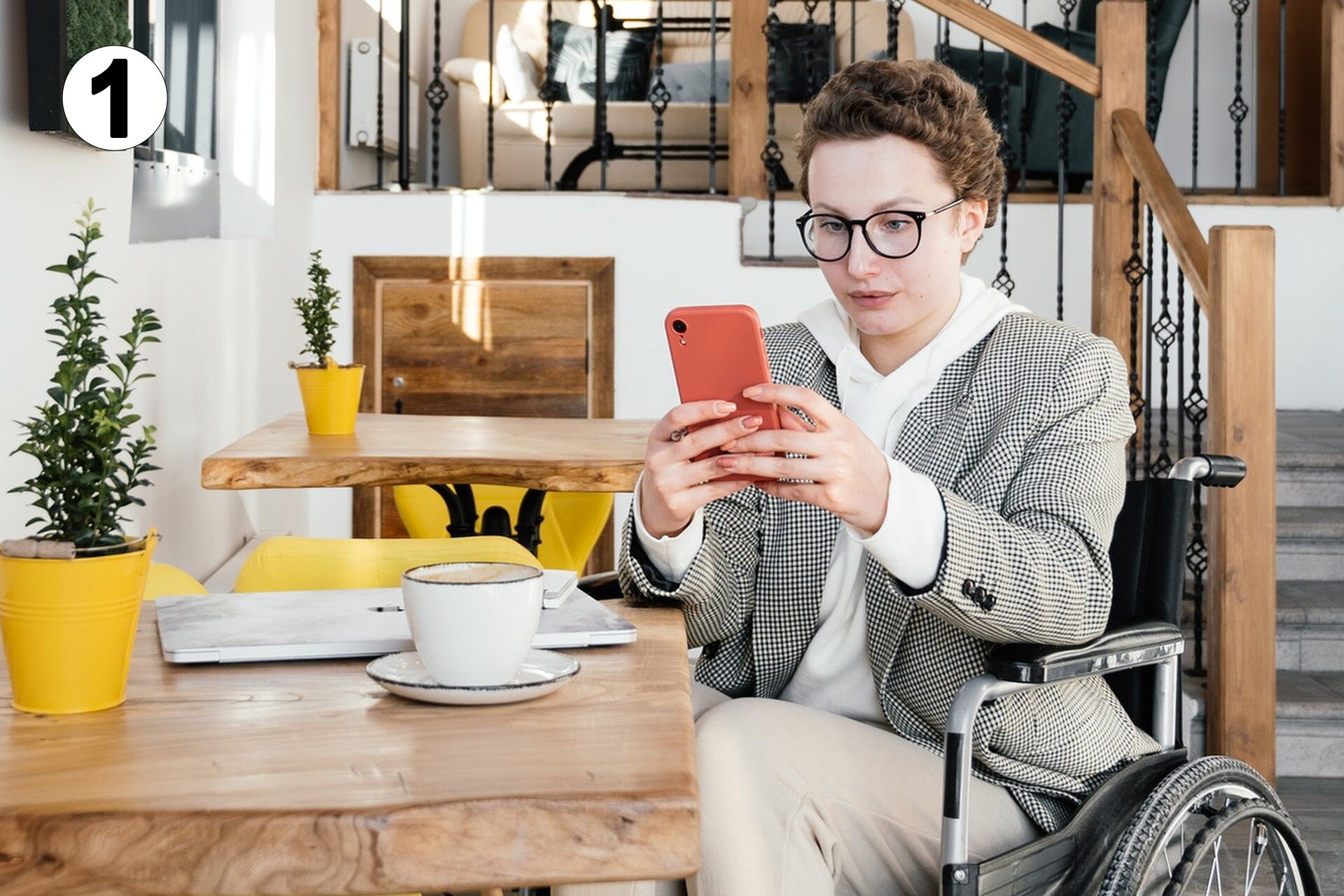 Pierwsze zdjęcie przedstawia kobietę w okularach, która siedzi przy stole, i wpatruje się w telefon. Kobieta znajduje się na wózku inwalidzkim. Na stole przed nią stoi filiżanka i leży zamknięty laptop.