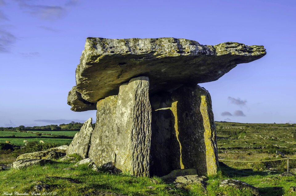 Fotografia przedstawia dolmen Poulnabrone w Burren w Irlandii. Składa się on z trzech pionowych chropowatych kamieni, na których ułożony jest jeden duży, płaski kamień leżący poziomo. Na kamieniach znajdują się ślady erozji. Niektóre z nich porośnięte są mchem. Dolmen usytuowany jest na zazielenionym, rozległymi pagórkowatym terenie. Na horyzoncie widać niskie drzewa. Niebo nad dolmenem jest błękitne, z delikatnymi białymi chmurami.