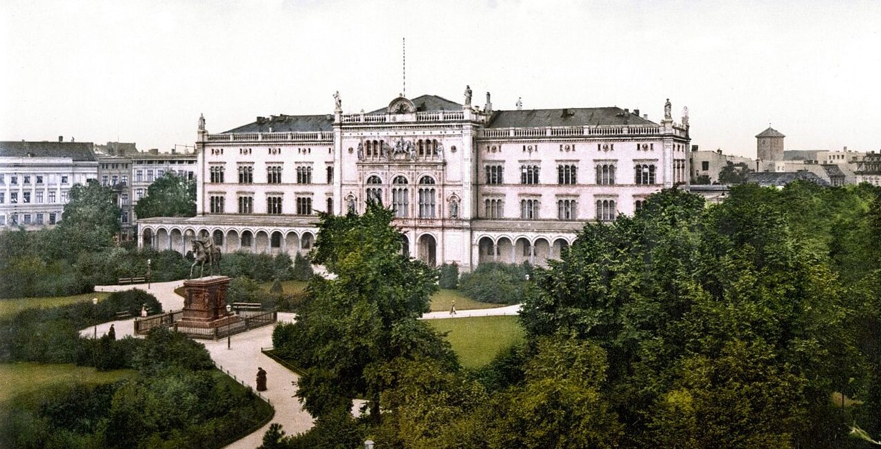 Ilustracja przedstawia Uniwersytet w Królewcu. Jest to biały budynek w starodawnym stylu z ozdobnymi fasadami, kolumnami i wieżyczkami. Wokół niego znajduje się park i alejki, na środku wielki pomnik mężczyzny na koniu.