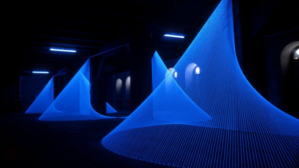 Na zdjęciu jest ukazane ciemne pomieszczenie z kilkoma instalacjami w kształcie stożka o kolorze niebieskim. Nad instalacją znajdują się 3 wąskie, podłużne lampy. W tle są łukowate okna.