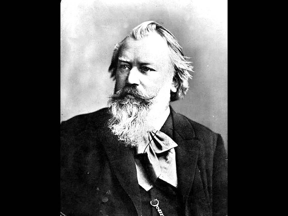 Ilustracja przedstawia Johannesa Brahmsa – niemieckiego kompozytora, pianistę i dyrygenta. Starszy mężczyzna z długą brodą i zakręconym wąsem. Twarz ma poważną. Ubrany jest w ciemny garnitur.