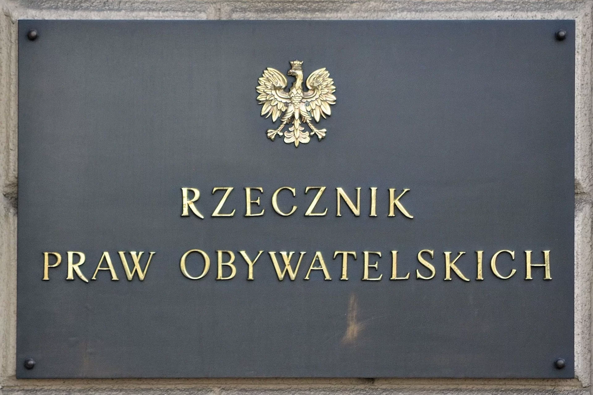 Zdjęcie przedstawia tablicę przymocowana do ściany. Widnieje na niej napis Rzecznik Praw Obywatelskich. Nad napisem znajduje się orzeł w koronie.