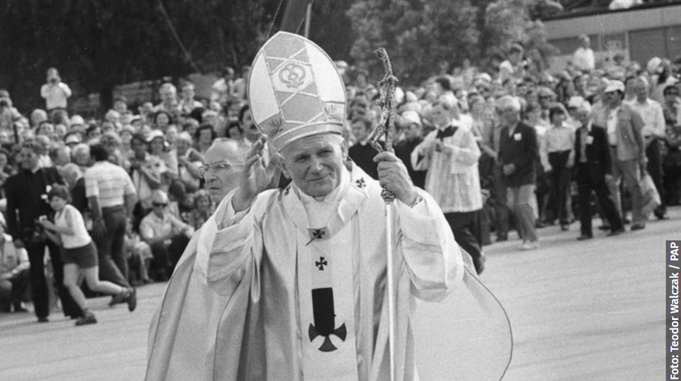 Zdjęcie przedstawia Jana Pawła II w papieskim stroju i tiarze na głowie, z pastorałem w dłoni. Papież błogosławi stojących przed nim ludzi.