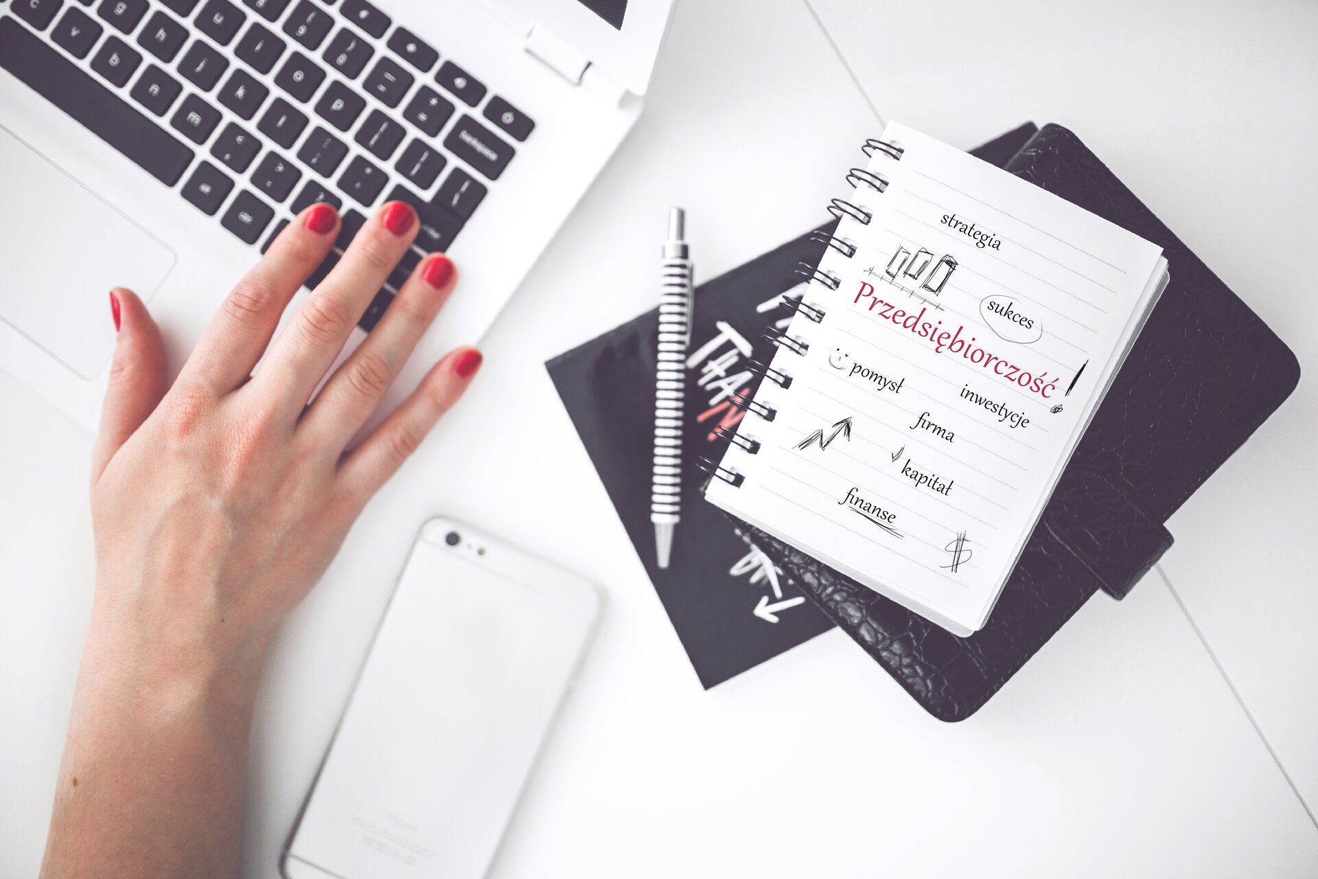 biurko, dłoń położona na klawiaturze laptopa, długopis, podkładka na której leży notatnik z napisem sukces, przedsiębiorczość, telefon komórkowy 