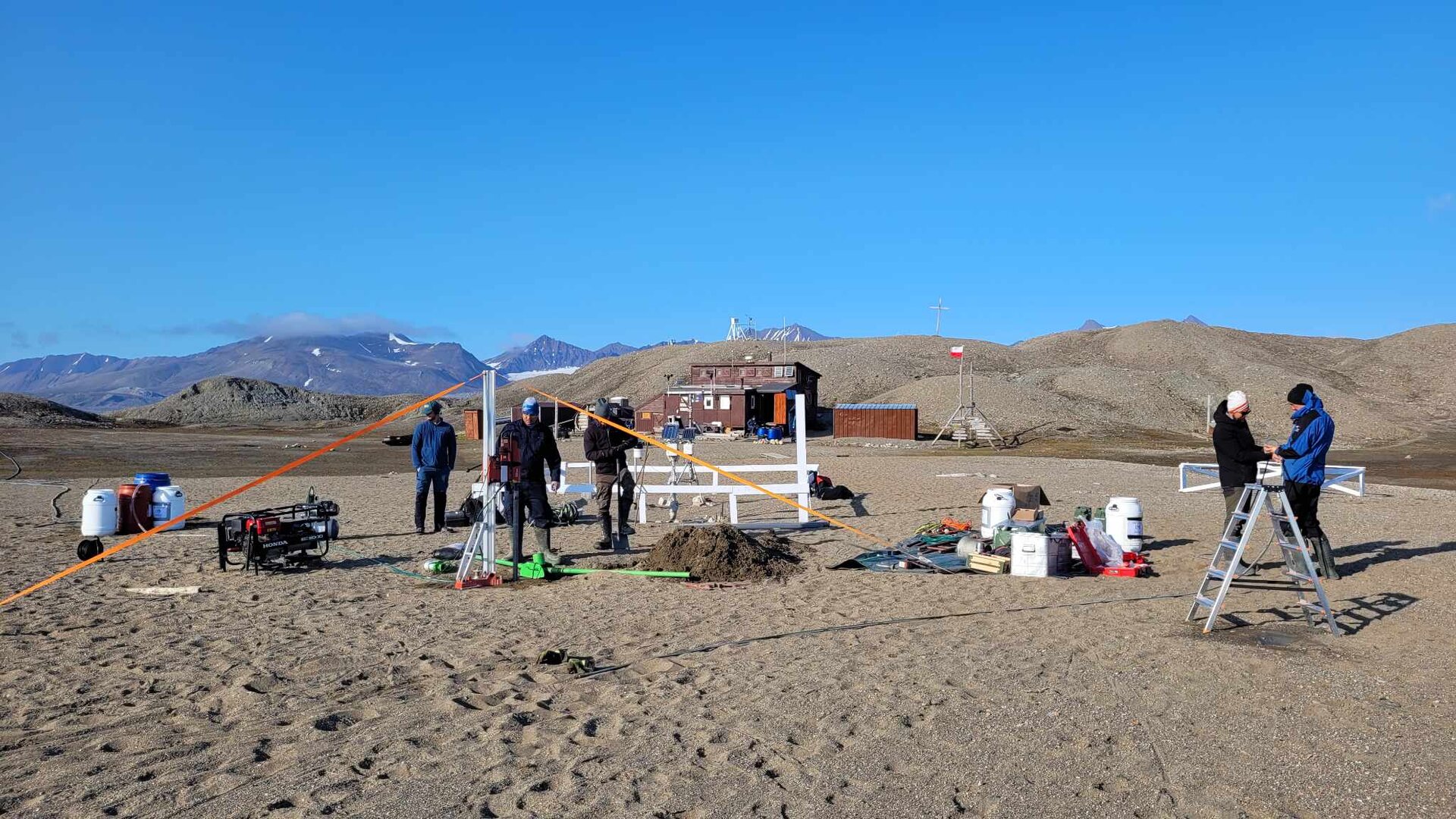 Na pierwszym planie zdjęcia grupa badaczy rozstawiających sprzęt pomiarowy, różne pojemniki, drabiny. Ubrani w ciepłą odzież. Dookoła jałowy teren, szary piasek, na drugim planie zabudowanie stacji badawczej i wzniesienia.