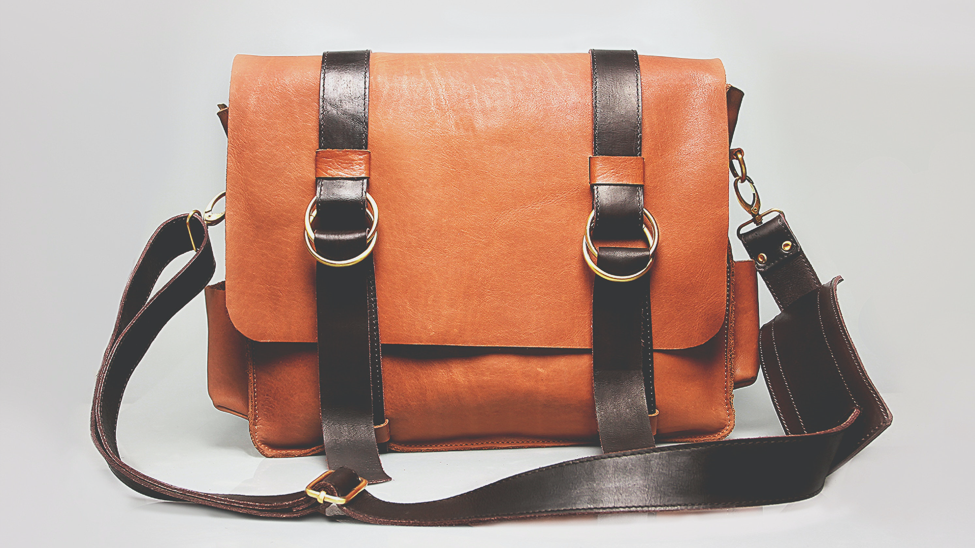 Zdjęcie przedstawia skórzaną torbę popularnie nazywaną listonoszką. Ma ona długi pasek służący do noszenia jej na ramieniu. Torba zapinana jest przez dwa mniejsze paseczki.