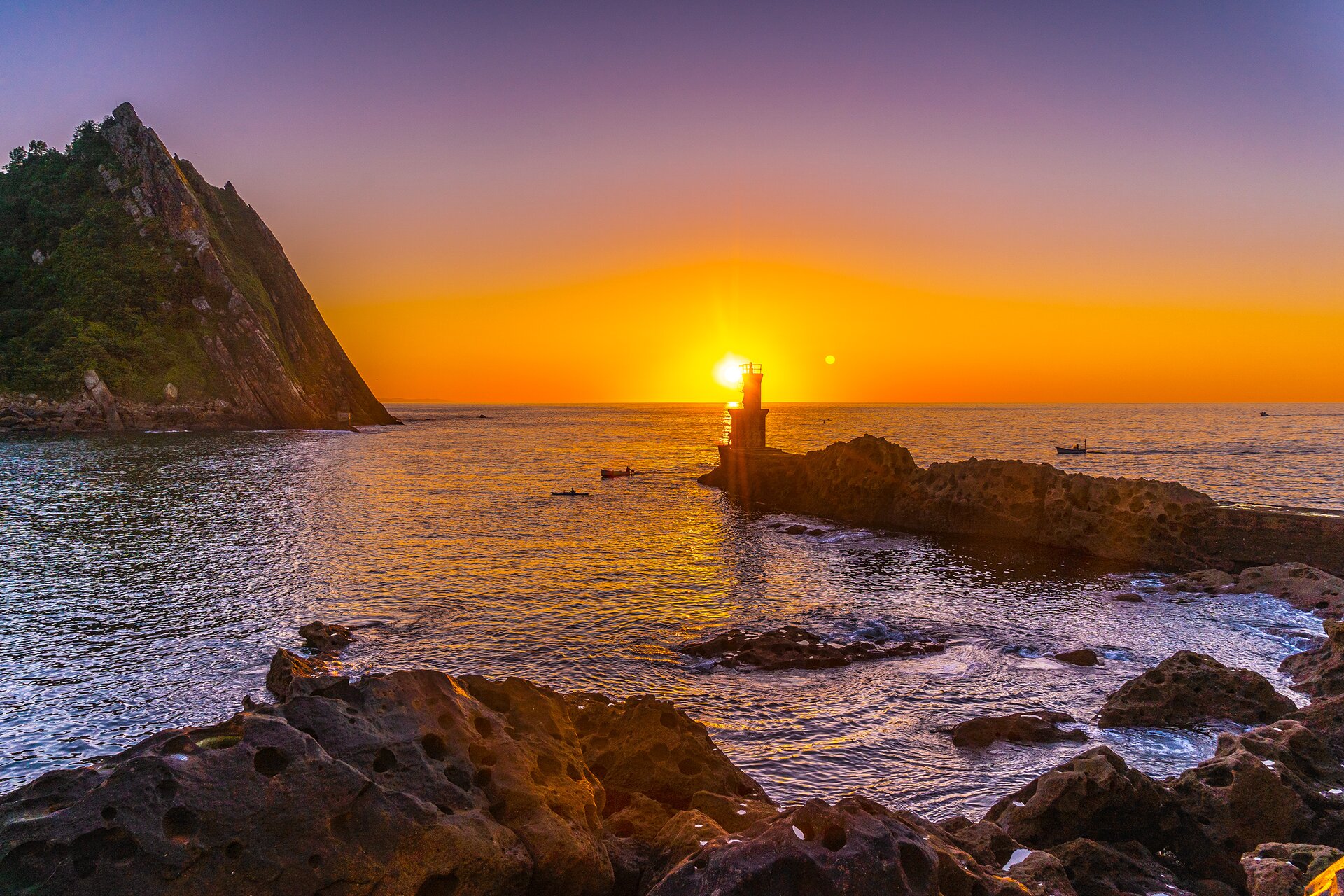 Zdjęcie przedstawia zachód słońca nad morzem. Widoczny jest zbiornik wodny ze spokojną, czarno‑fioletową wodą, otoczony skalistym wybrzeżem. Po prawej stronie widoczny jest cypel, na końcu którego  znajduje się niewielka latarnia morska. Poniżej widoczne są dwie niewielkie łódki po lewej i jedna po prawej stronie cypla. W lewej, górnej części widoczne jest wysokie, skaliste wyniesienie. Promienie zachodzącego słońca oświetlają krajobraz i nadają mu pomarańczową tonację. 
