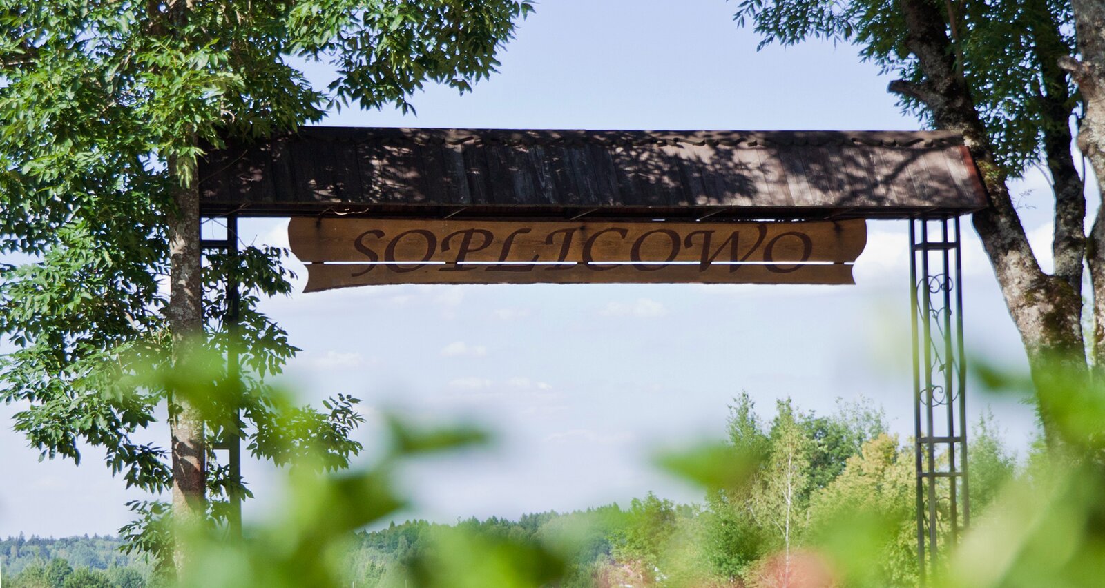 Zdjęcie przedstawia drewnianą bramę, na której nad wjazdem umieszczono drewniany daszek, a pod nim zawieszono tablicę z napisem „SOPLICOWO". Z obu stron bramy widoczne są wysokie, zielone drzewa. W tle widoczny jest fragment lasu oraz błękitne niebo. Zdjęcie zostało wykonane z pewnej odległości, z dolnej perspektywy – widoczne są zamazane fragmenty roślinności u dołu zdjęcia.