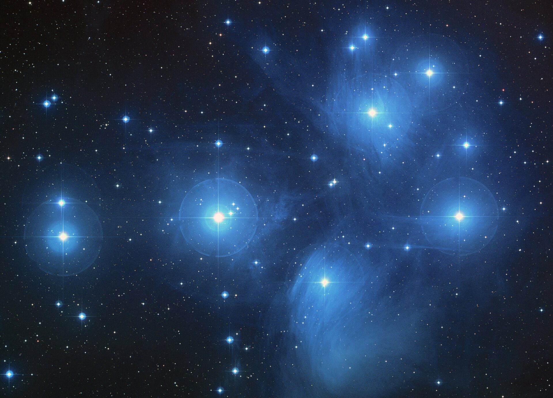 Grafika przedstawia gromadę gwiazd Plejady w Gwiazdozbiorze Byka. Tło czarne. Na grafice widoczne mnóstwo jasnoniebieskich punktów różnej wielkości. Kilka największych wydają się być otoczone niebieską otoczką, łuną.