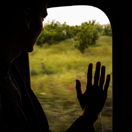 Fotografia przedstawia osobę wyglądającą przez okno jadącego pociągu, trzymając dłoń na szybie.