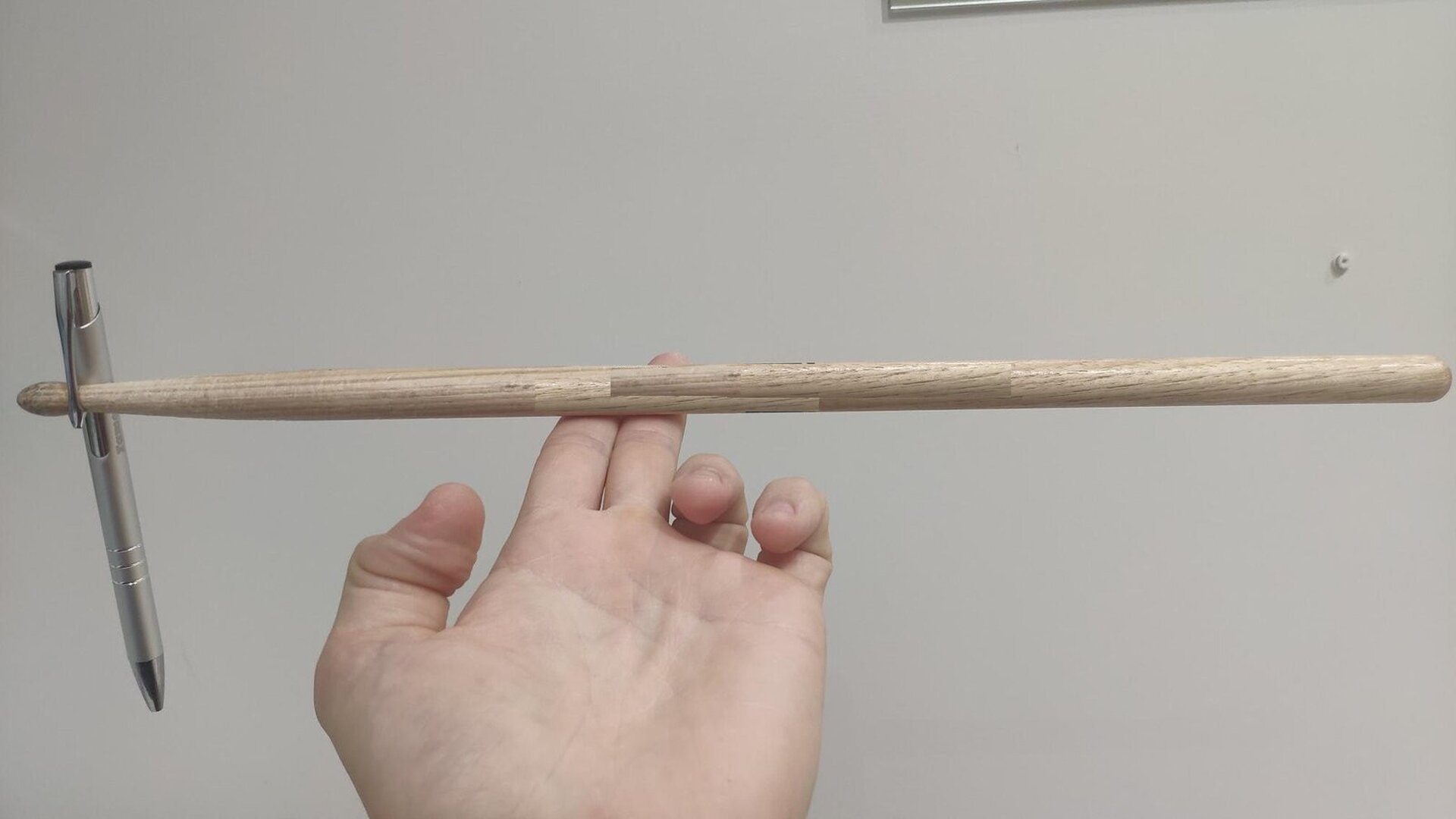 Fot. 2. Zdjęcie przedstawia pałeczkę perkusyjną opartą na wyprostowanych dwóch palcach (wskazującym i środkowym) na ułożonej poziomo dłoni. Do lewego końca pałeczki przyczepiony jest długopis. Pałeczka podparta w punkcie leżącym na lewo od środka.