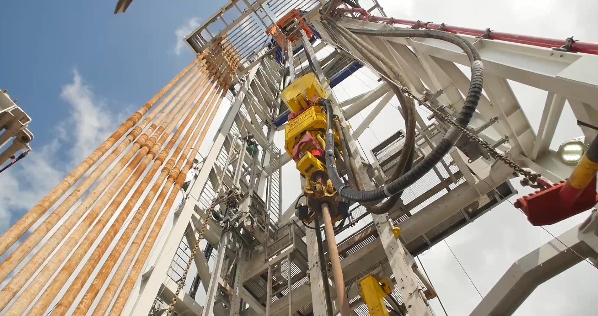 Na zdjęciu widoczny jest fragment wieżomasztu widoczny od dołu. W centralnej części zdjęcia znajduje się napęd Top Drive. Od dolnej części napędu odchodzi podłużna cylindryczna rura. Napęd znajduje się pomiędzy stalową konstrukcją kratową o kształcie podłużnego prostopadłościanu o orientacji pionowej. Po lewej stronie napędu widocznych jest dziesięć rur. Rury są magazynowanie równolegle względem siebie i względem wieży. Po prawej stornie wieży zamontowane są dwa czarne węże które łączą się z napędem.