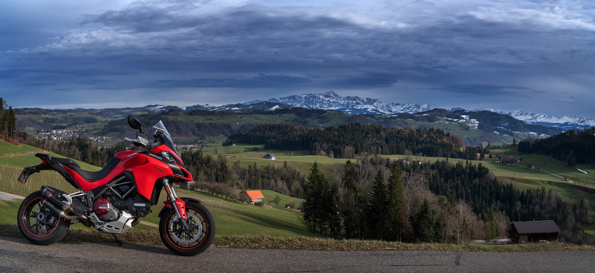 Zdjęcie przedstawia motocykl stojący na szutrowej drodze w terenie pagórkowatym. Tył motocykla znajduje się znacznie ponad tylnym kołem. Znaczna część motocykla posiada osłony.  