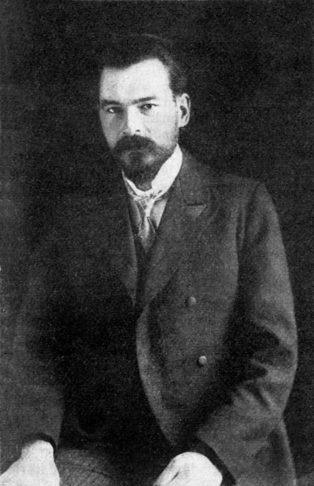 Czarno białe zdjęcie przedstawia polskiego fizyka Mariana Smoluchowskiego. Mężczyzna w wieku około trzydziestu lat. Blada skóra twarzy gładko ogolona. Czoło wysokie. Widoczne zakola. Czarne gęste włosy zaczesane do tyłu. Duże widoczne uszy. Brwi ciemne, gęste. Oczy małe. Nos duży. Długie gęste wąsy sięgają dolnej części policzków. Dolna szczęka z niewielkim ciemnym krótkim zarostem. Mężczyzna ubrany w białą koszulę i długą ciemną marynarkę. Dłonie oparte na kolanach.