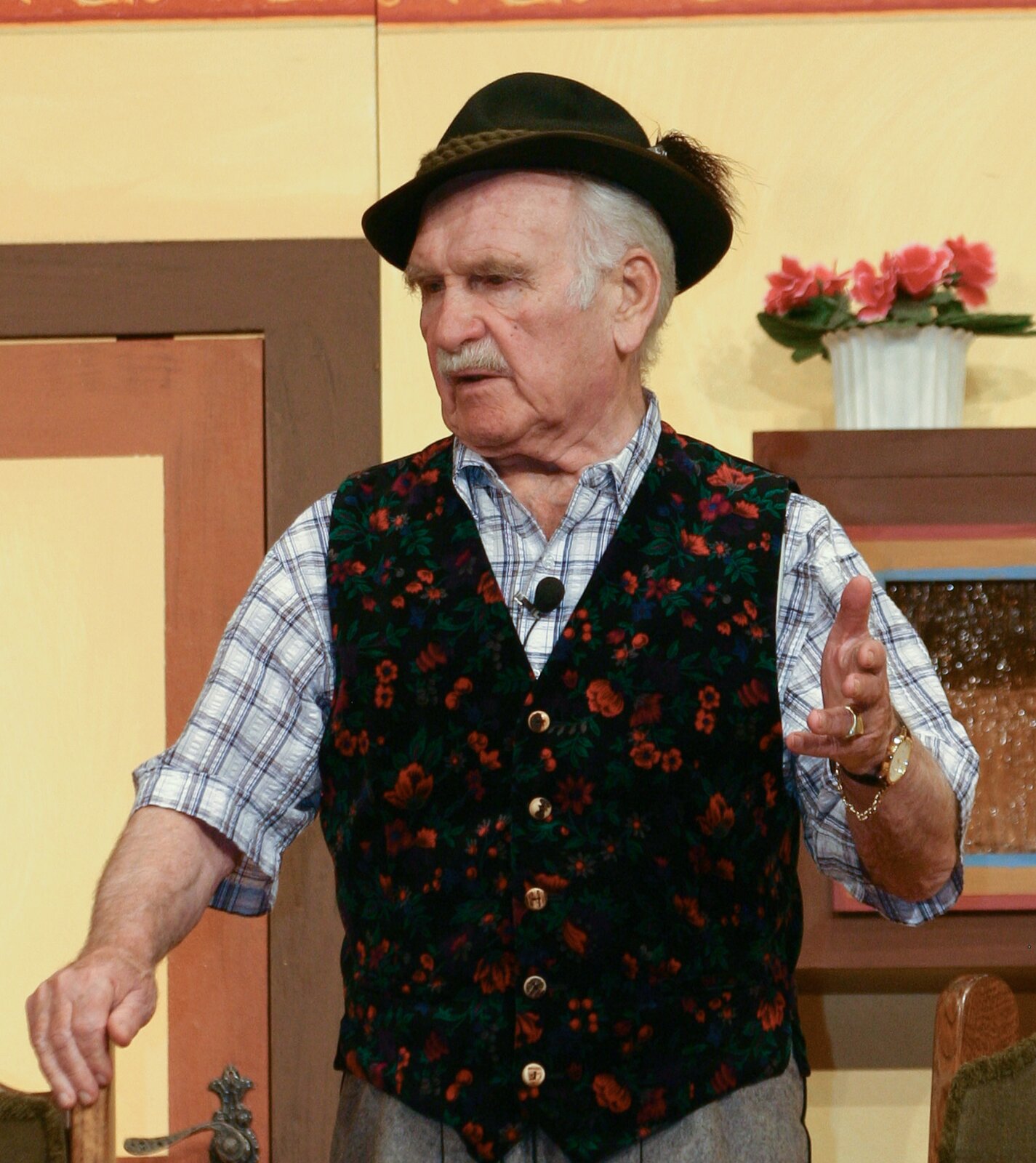 Zdjęcie przedstawia przykładowy plan średni. Widoczny jest starszy, stojący mężczyzna, pokazany od pasa w górę. Ma rozłożone ręce, gestykuluje, jego głowa jest lekko zwrócona w lewo. Ubrany jest w kraciastą koszulę, kamizelkę w kwiaty na czarnym tle, kapelusz z piórkiem. W tle widoczne są drzwi i mebel.