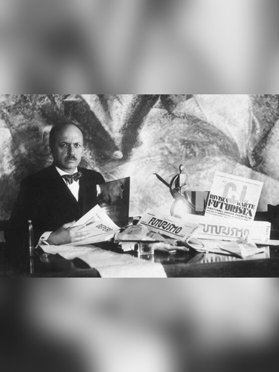 Zdjęcie przedstawia dojrzałego mężczyznę w garniturze, koszuli z muszką. Mężczyzna siedzi przy biurku, na którym leży wiele porozrzucanych egzemplarzy gazet.