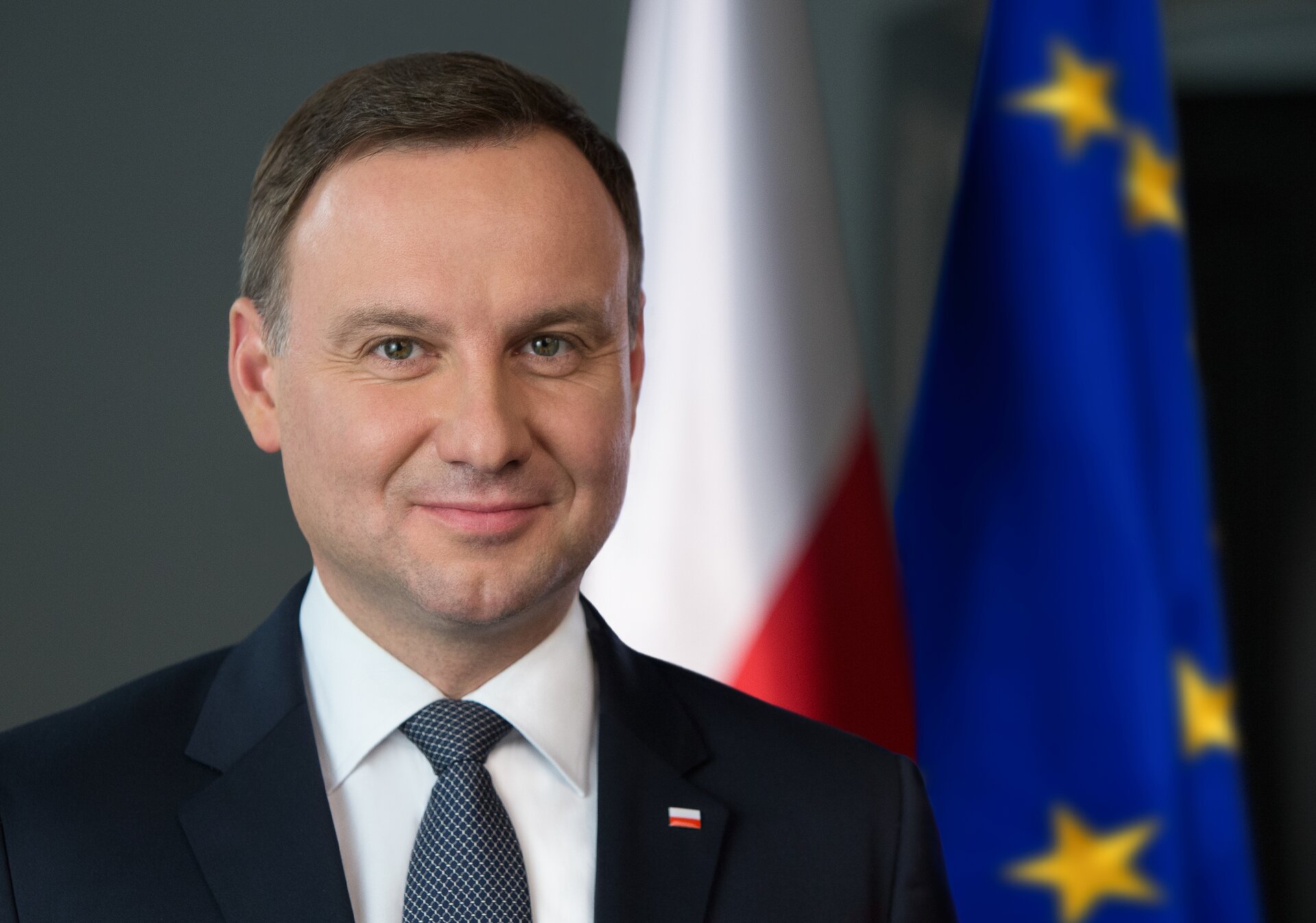 Fotografia portretowa przedstawiająca prezydenta Andrzeja Dudę.na tle flagi Rzeczypospolitej Polskiej oraz Unii Europejskiej. Mężczyzna o okrągłej twarzy, z krótkimi, ciemnymi włosami zaczesanymi na prawą stronę. Uśmiecha się. Ubrany w koszule, krawat i marynarkę. W klapie marynarki znaczek z flagą Polski.