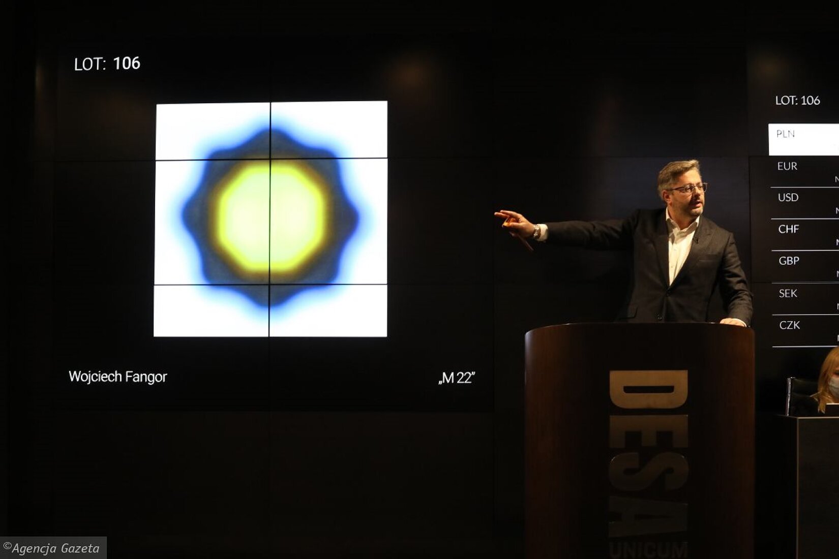 Na zdjęciu po prawej stronie znajduje się mężczyzna, jego prawa ręka wskazuje na obraz, a głowa zwrócona jest w przeciwnym kierunku. Obraz ukazuje trzy rozety, które są ze sobą połączone. Rozety są koloru żółtego, ciemnożółtego następnie przechodzą w kolor niebieski. Nad obrazem, po lewej stronie znajduje się napis: „LOT: 106”.  Poniżej widnieje biały podpis: „Wojciech Fangor”. Tło zdjęcia jest czarne.  