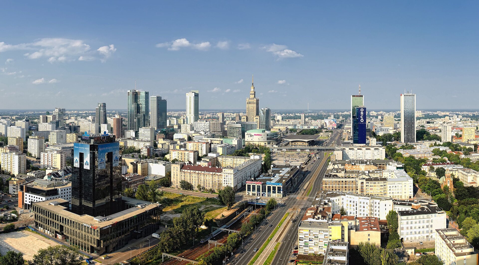 Zdjęcie przedstawia widok z góry na centrum Warszawy. Na pierwszym planie szeroka, kilkupasmowa ulica. Wokół kamienice, bloki i wieżowce.