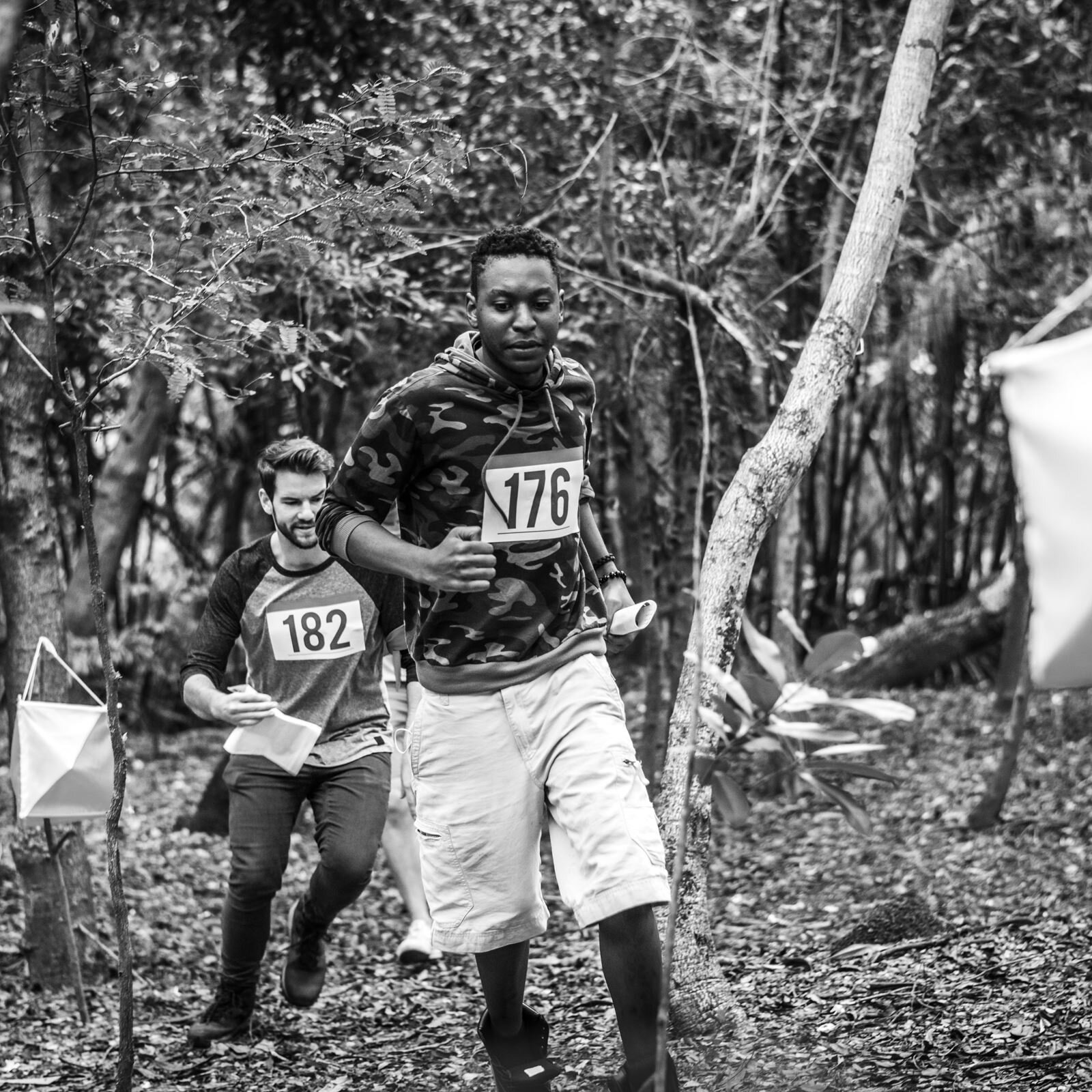 Czarno‑białe zdjęcie przedstawia dwóch młodych mężczyzn biegnących w lesie. Mężczyzna na pierwszym planie ma krótkie, ciemne włosy, jest ubrany w bluzę z kapturem i jasne szorty. Na piersi ma numer 176. Za nim biegnie drugi mężczyzna, który ma jasne, krótkie włosy, ubrany jest w bluzę z numerem 182 na piersi oraz długie spodnie. Obydwaj trzymają w ręku kartki. Po lewej stronie widoczny jest sześcienny jasny znacznik umieszczony na drzewie.