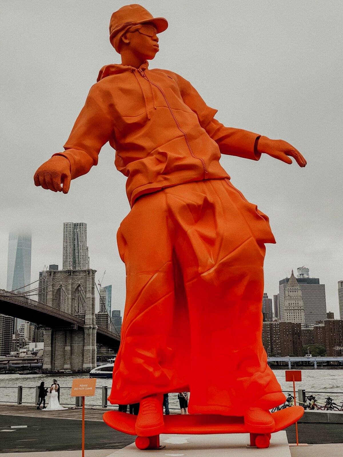 Zdjęcie przedstawia nowoczesną rzeźbę Skate'a ustawioną na nadrzecznej promenadzie w wielkim mieście. Rzeźba wykonana jest ze sztucznego tworzywa i pokryta jednakowym ciepłym kolorem, pomarańczowym. Przedstawia chłopaka na deskorolce, który ma na sobie czapkę z daszkiem, okulary, luźną kurtkę i szerokie spodnie. Rzeźba stara się upodobnić do autentycznej postaci, wyróżnia się z daleka, wprowadza element zabawy. 