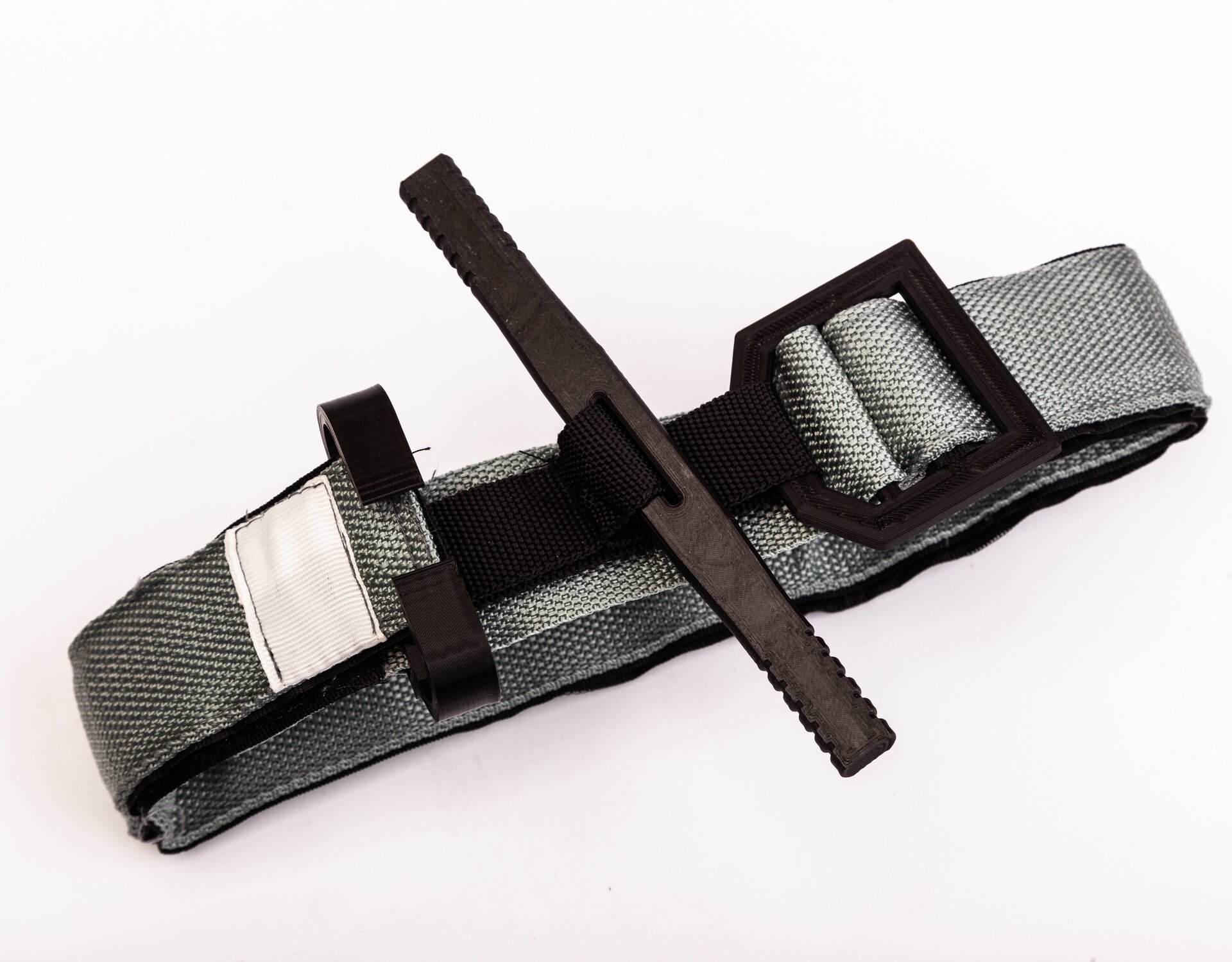 Zdjęcie przedstawia szarą opaskę uciskową - stazę taktyczną z czarnymi elementami. Po środku opaski znajduje się czarny pręcik umiejscowiony prostopadle do niej.