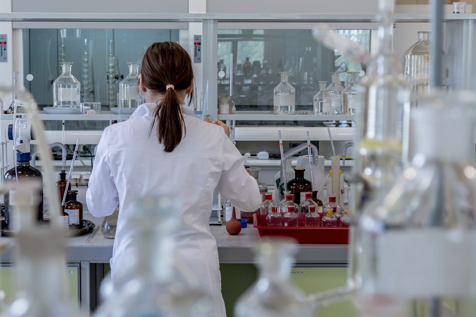 Zdjęcie przedstawia kobietę w białym fartuchu, która pracuje w laboratorium. Jest odwrócona tyłem do fotografa. Na blacie znajdującą się przed ną znajdują się liczne tacki, butelki, menzurki i probówki.