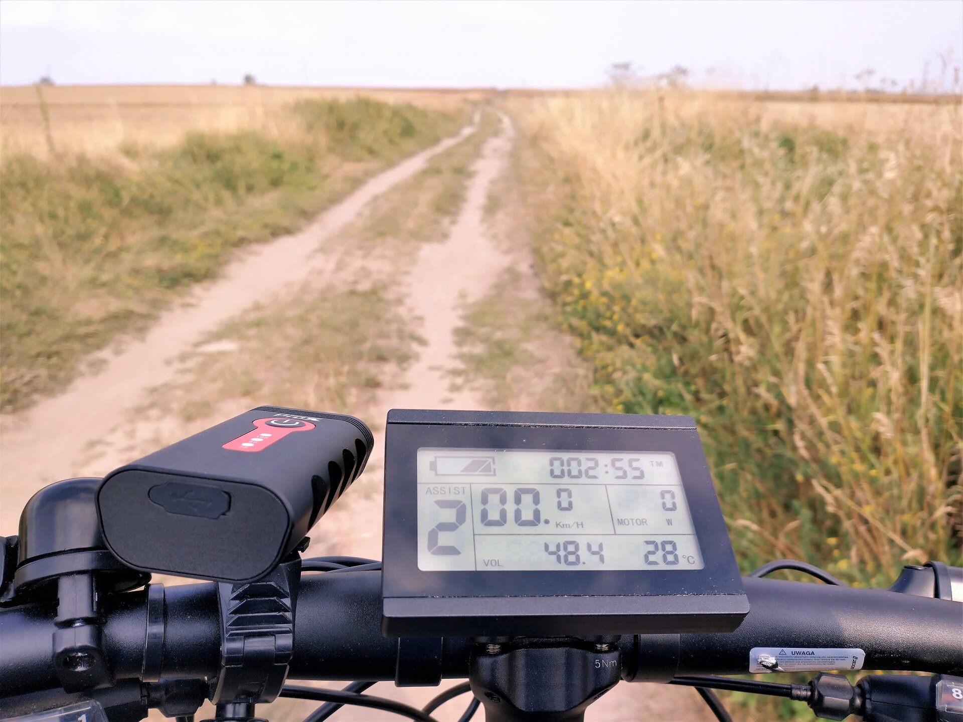 Rys. a. Ilustracja przedstawia zdjęcie licznika rowerowego. Licznik rowerowy posiada prostokątny wyświetlacz ciekłokrystaliczny, na którym wyświetlana jest aktualna prędkość zero kilometrów na godzinę z dokładnością do jednej dziesiątej kilometra na godzinę. Poniżej aktualnej prędkości widoczna jest informacja o aktualnym przebiegu od ostatniego wyzerowania licznika równa czterdzieści osiem przecinek cztery kilometrów. Następnie po prawej wyświetla się aktualna temperatura dwadzieścia osiem stopni celsjusza. Na samej górze widać stan naładowania baterii oraz czas przejazdu równy dwie godziny i pięćdziesiąt pięć minut. Wokół wyświetlacza widoczna jest czarna, plastikowa ramka.