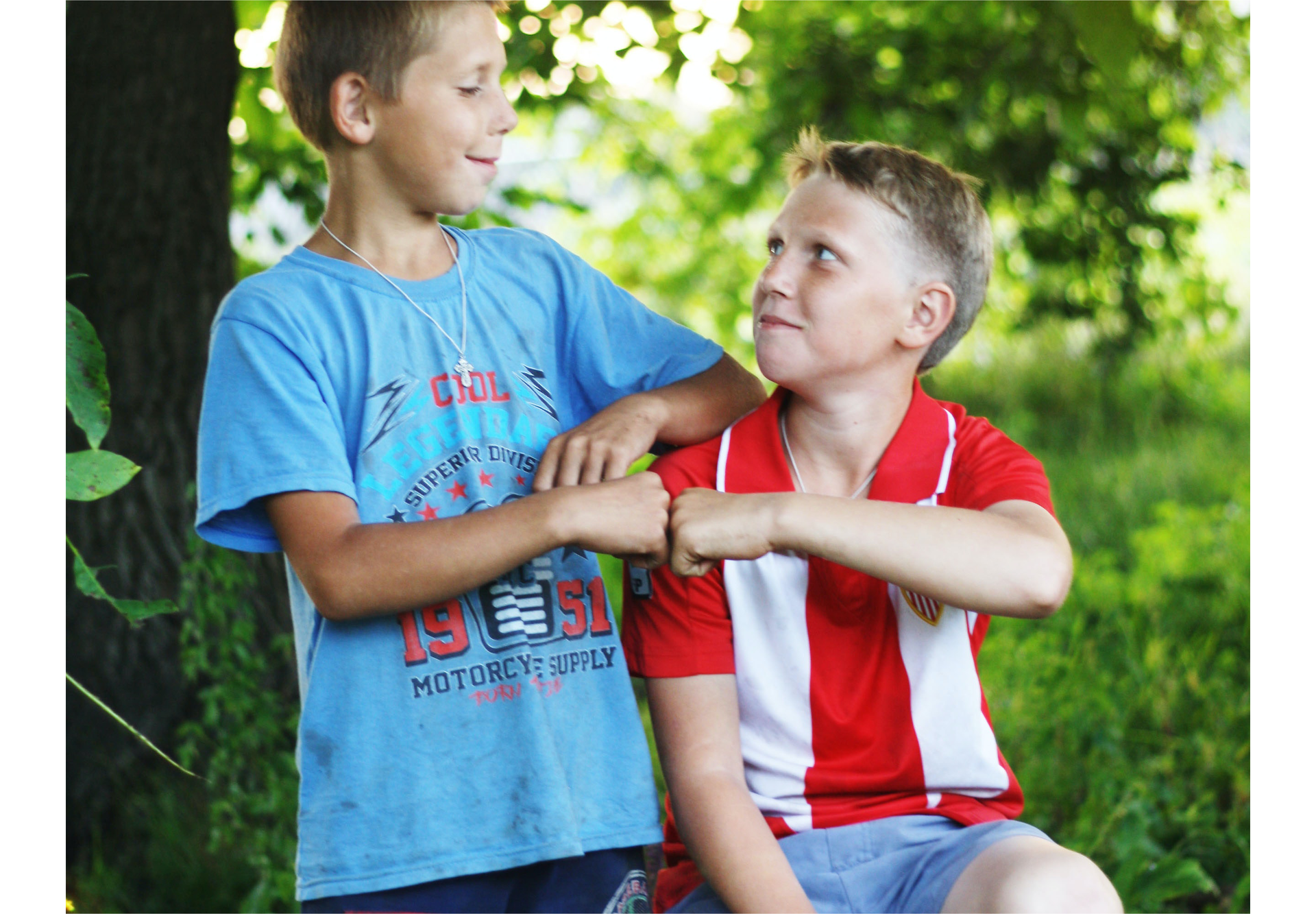 Zdjęcie przedstawia dwóch nastoletnich chłopców na tle drzew. Chłopiec po lewej stronie stoi. Chłopiec po prawej stronie siedzi. Chłopcy witają się popularnym „żółwikiem”, czyli dotykają się zaciśniętymi pięściami. 