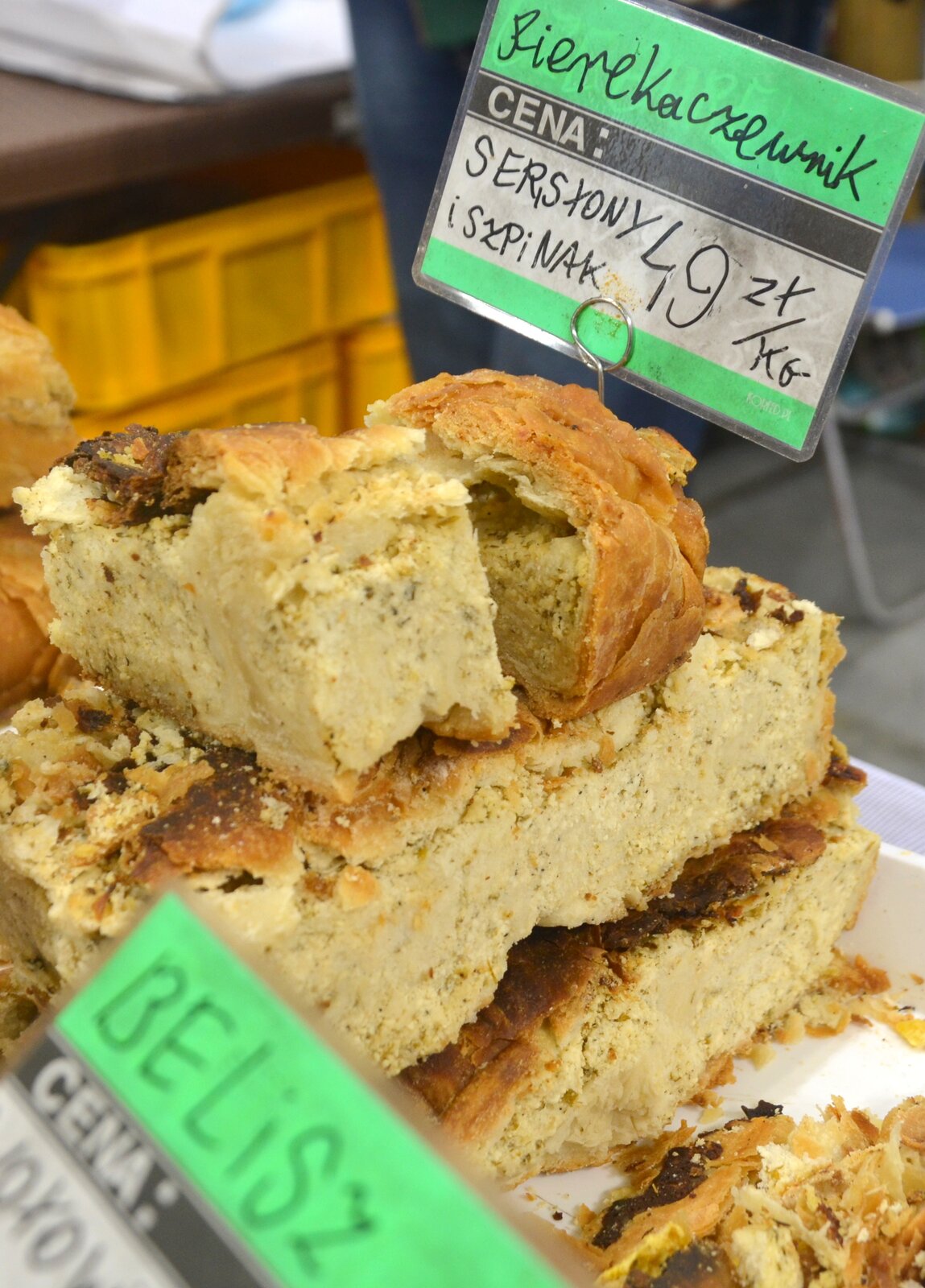 Na zdjęciu znajdują się pierekaczewniki. Jest to potrawa w formie zapiekanego ciasta z nadzieniem. Ciasto ułożone jest w kawałkach, jeden na drugim, a na górze znajduje się cena za kilogram. 