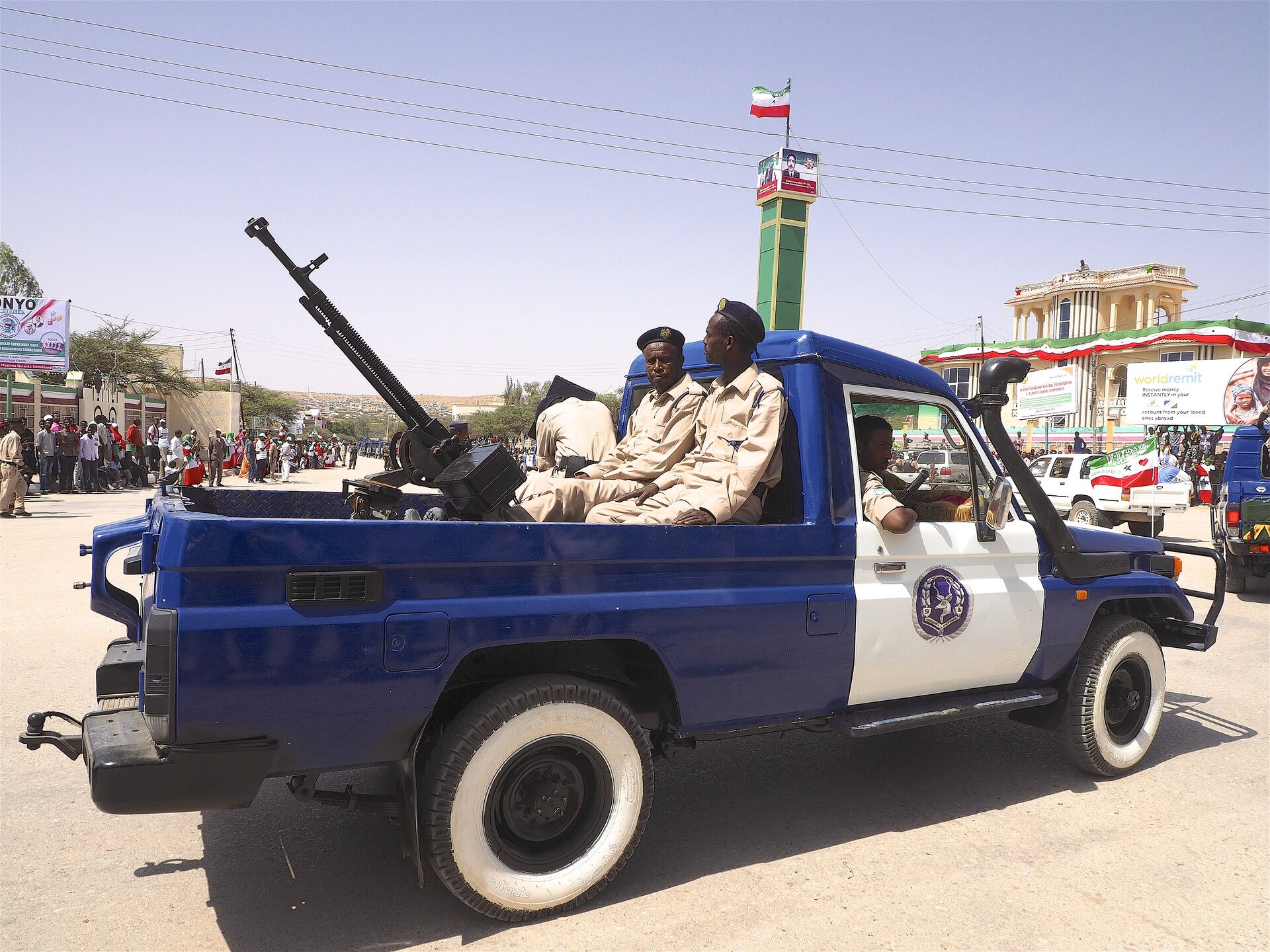Zdjęcie przedstawia siły zbrojne Somalilandu. Na pace ciężarówki siedzi dwóch ciemnoskórych mężczyzn w jasnobrązowych mundurach. Przed nimi jest karabin. Pojazd jest niebieski z białymi drzwiami. W tle pod budynkami stoi tłum ludzi. Budynki ozdobione są flagami.