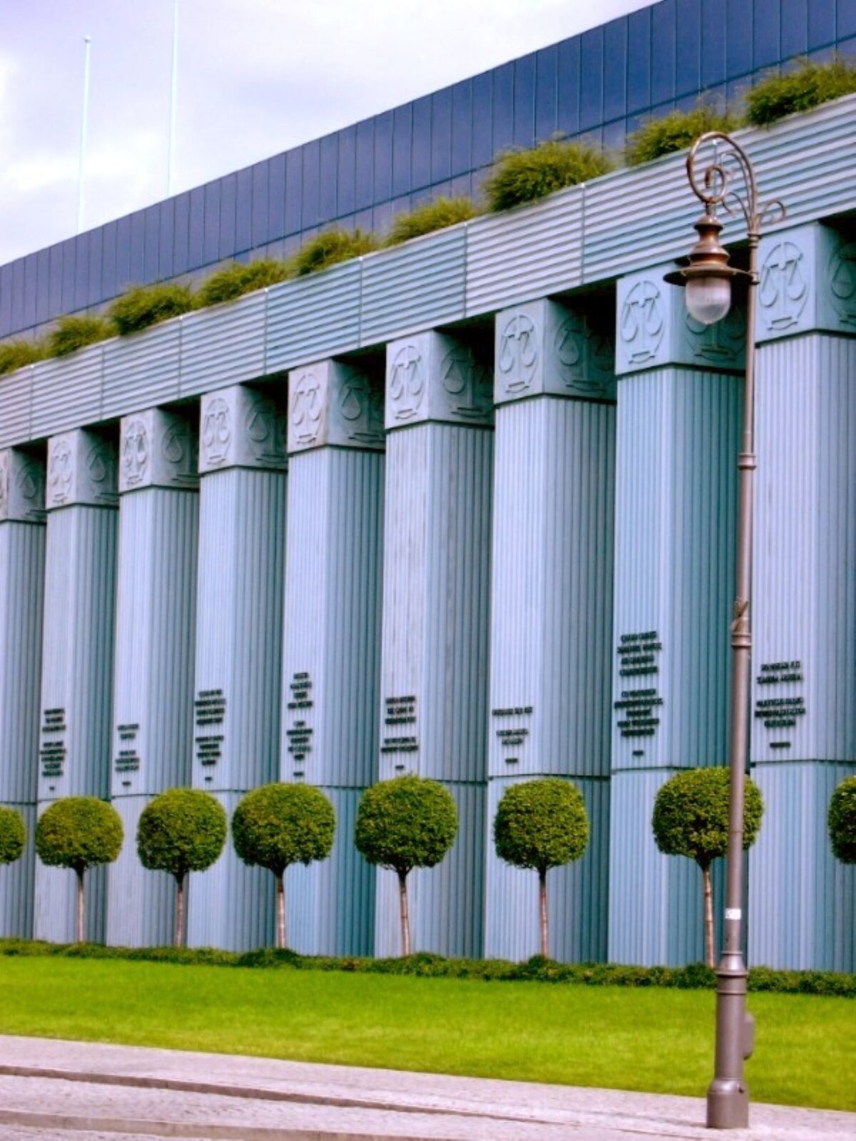 Ilustracja przedstawia fotografię „Paremie prawnicze na kolumnach gmachu Sądu Najwyższego”. Na zdjęciu znajdują się wysokie, szare kolumny. Na ich szczycie są płaskorzeźby przedstawiające wagi, a w dolnej części czarne napisy. Nad kolumnami znajduje się zielona roślinność, a wyżej jest grafitowa ściana. Przed budynkiem jest jasnozielony trawnik i niskie drzewa liściaste. 