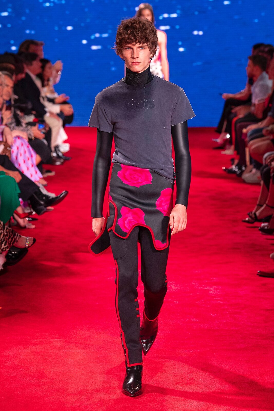 Fotografia przedstawia modela na wybiegu. Ma on na sobie szarą koszulkę z krótkim rękawem, spod której widoczne są skórzane rękawy oraz golf; czarne spodnie z czerwonymi liniami po bokach oraz lakierowane buty ze spiczastymi czubkami. Pomiędzy jego bluzką a spodniami widoczny jest czarny element materiału w różowe róże. W tle widać  siedzących ludzi. Dywan, po którym chodzi model, jest czerwony.
