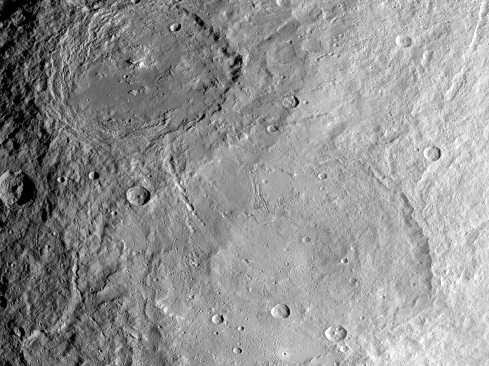 Rys. 2. Zdjęcie poglądowe przedstawia dwa olbrzymie kratery Ceres uformowane w różnym czasie. W górnej części zdjęcia widoczny jest krater Urvara, który powstał około 120–140 mln lat temu. W dolnej części zdjęcia rysuje się znacznie mniej wyraźny krater Yalode, który jest starszy o prawie miliard lat. Yalode, ze względu na swój wiek, ma zatarte brzegi, jego struktura jest pokryta znacznie mniejszymi i młodszymi kraterami. Yalode powinien być znacznie głębszy niż jest teraz. Prawdopodobnie reakcje wewnętrznej aktywności sprawiły, że część skorupy w tym miejscu została wypiętrzona w górę zmniejszając głębokość krateru.