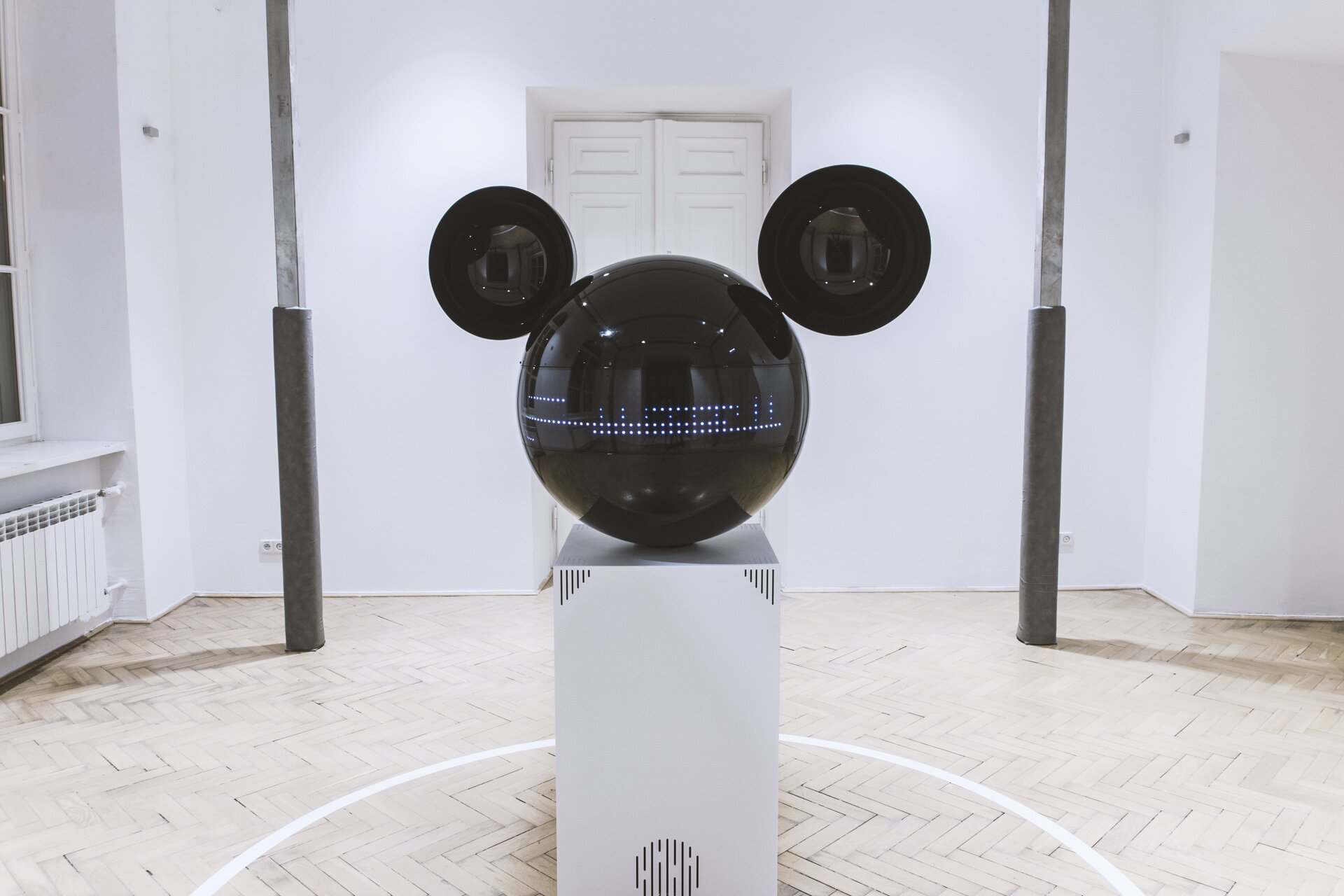 Zdjęcie przedstawia Mikifon. Jest to współczesna rzeźba w kształcie głowy Myszki Miki, składająca się z czarnej, błyszczącej kuli, dwóch wklęsłych półkul jako uszy oraz białej, wysokiej kolumny, na której stoi. Na głowie wyświetla się wizualizacja muzyki złożona z białych kropek. Na kolumnie umieszczono głośnik. Rzeźba znajduje się na drewnianej podłodze. Za nią stoją dwa słupy. Ściany są białe.