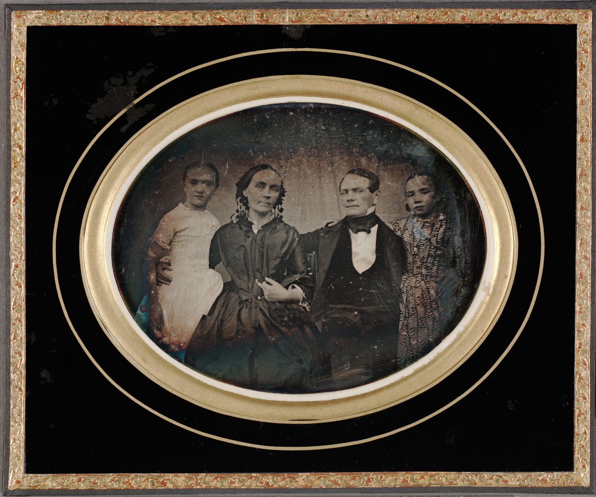 Zdjęcie przedstawia portret rodziny w okrągłej ramie. Na zdjęciu znajdują się dorosła kobieta i mężczyzna oraz dwie małe dziewczynki. Kobiety ubrane są w sukienki, mężczyzna ma na sobie garnitur.